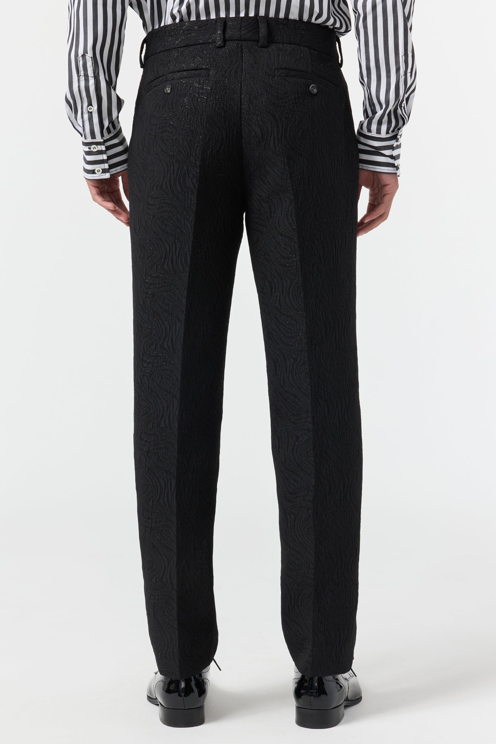 A person faces away, wearing Clyde Slim Trouser - Metallic Groove, a black belt, shiny black shoes, and a black-and-white striped long-sleeve shirt against a plain white background.