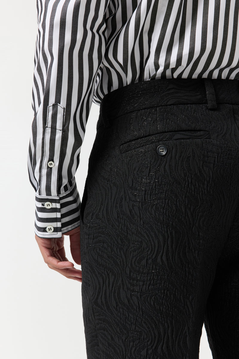 A person is shown from the waist down wearing the Clyde Slim Trouser - Metallic Groove, styled with a black and white vertically striped shirt and their left arm relaxed by their side.