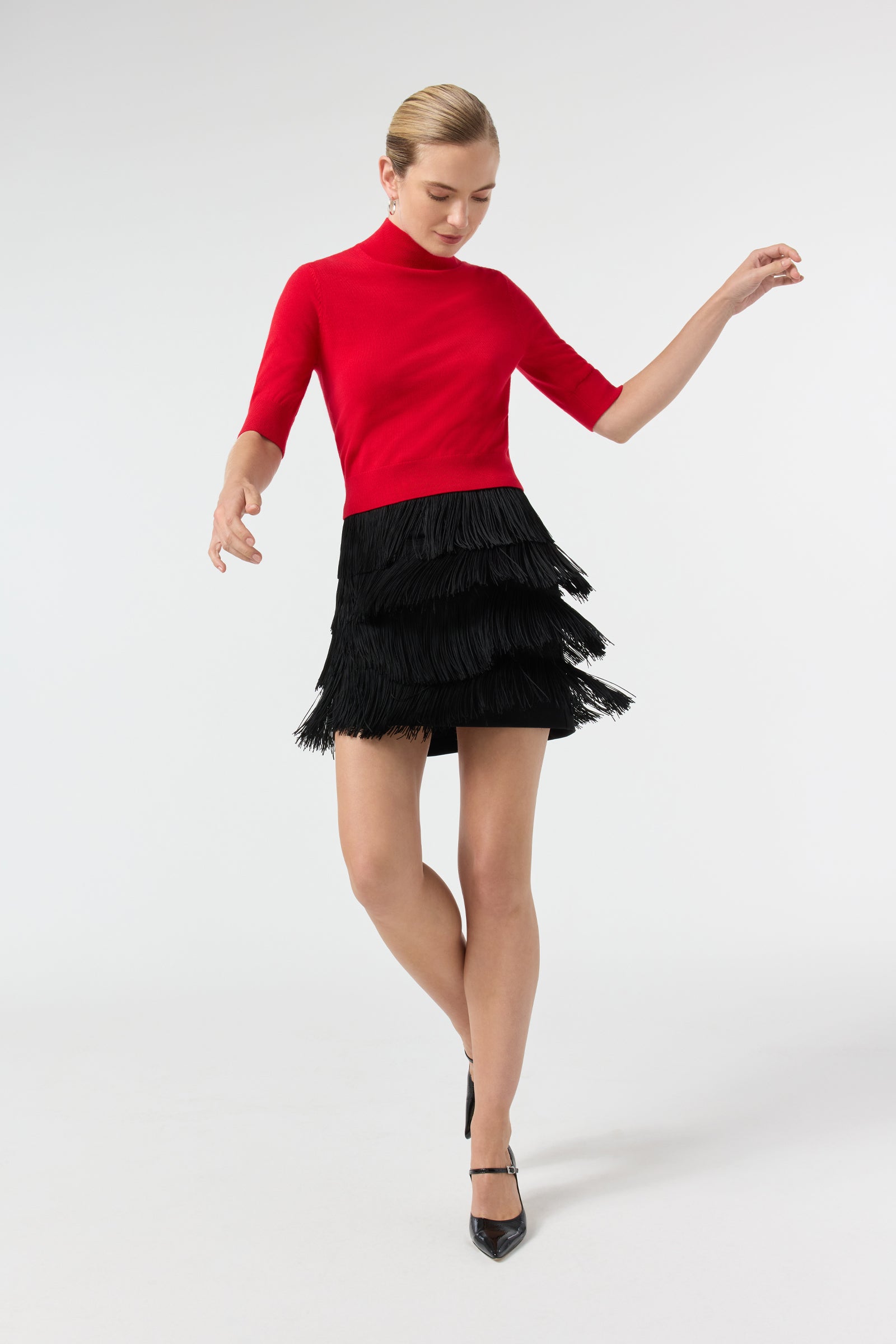 A woman models the Mali Mock Neck Pullover, a fitted red top with elbow-length sleeves, paired with a black fringe mini skirt as she poses gracefully against a plain white background, arms raised and one leg crossed.