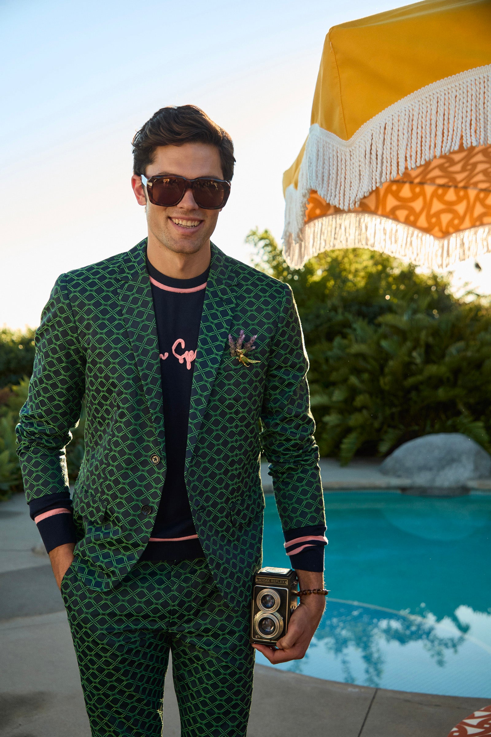 A man wears the Thurston Blazer in Clover Geo Jacquard and sunglasses as he smiles by a pool with a vintage camera. A yellow fringed umbrella and lush greenery can be seen in the background.