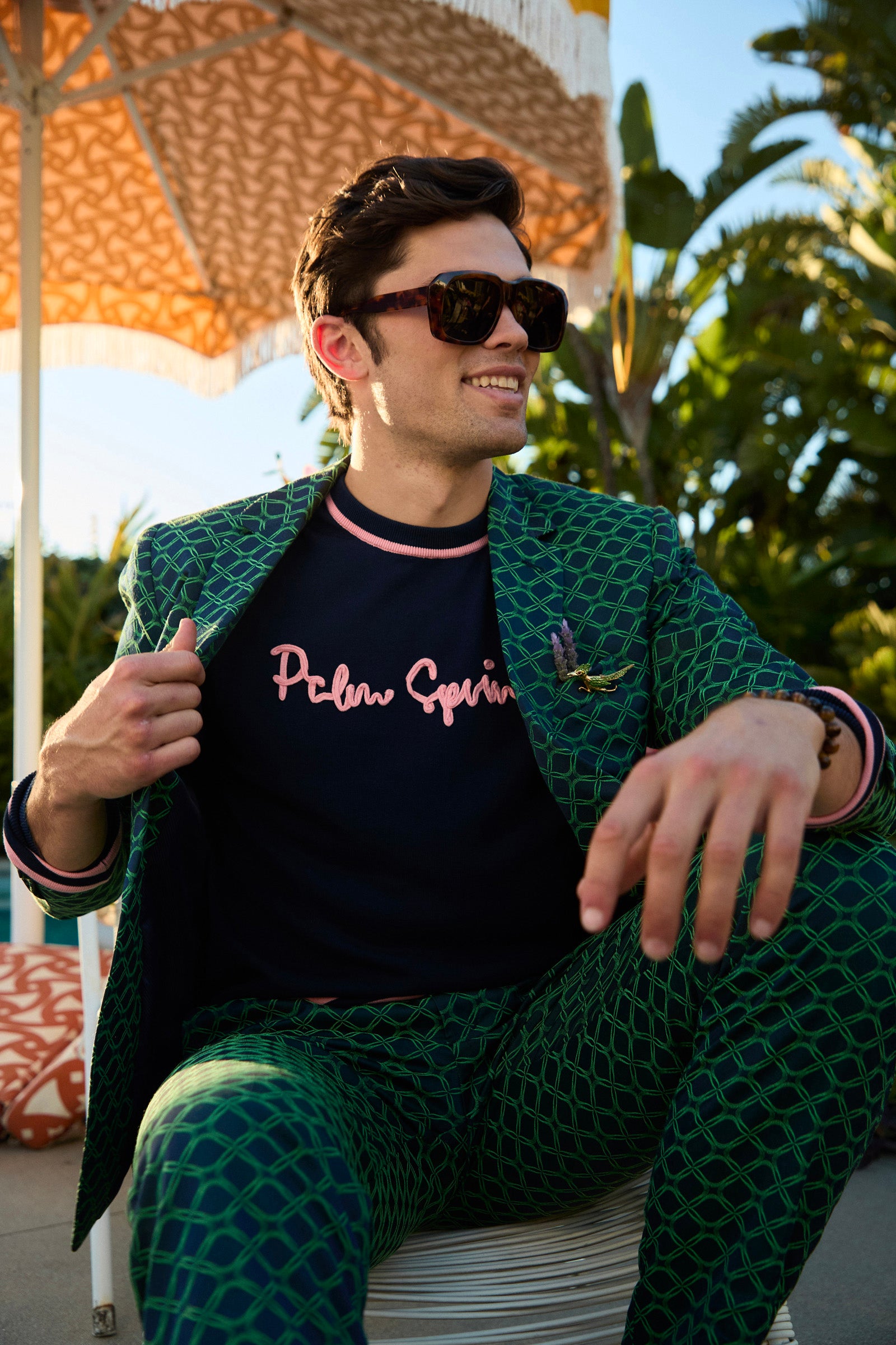 Man wearing sunglasses and a patterned outfit with 'Palm Springs' text, sitting outdoors.