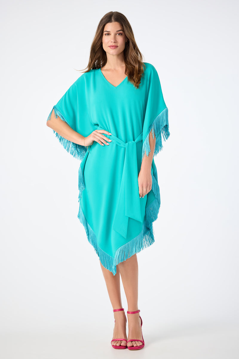 A woman with long brown hair and a neutral expression stands facing forward in the Toulon Teal Toulouse Short-Sleeve Fringe Dress, belted at the waist and paired with pink strappy high heels against a white background.