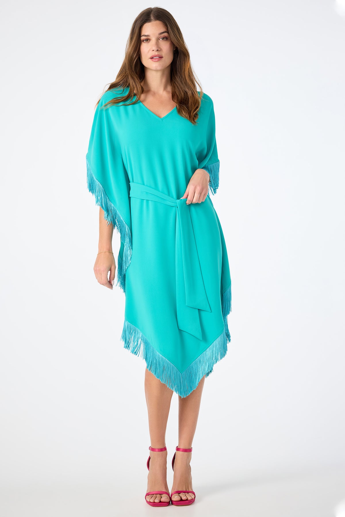 A woman models the Toulouse Short-Sleeve Fringe Dress, a loose turquoise style with fringe on the sleeves and hem, plus a matching waist tie. She pairs it with pink strappy heels and poses against a plain white background.
