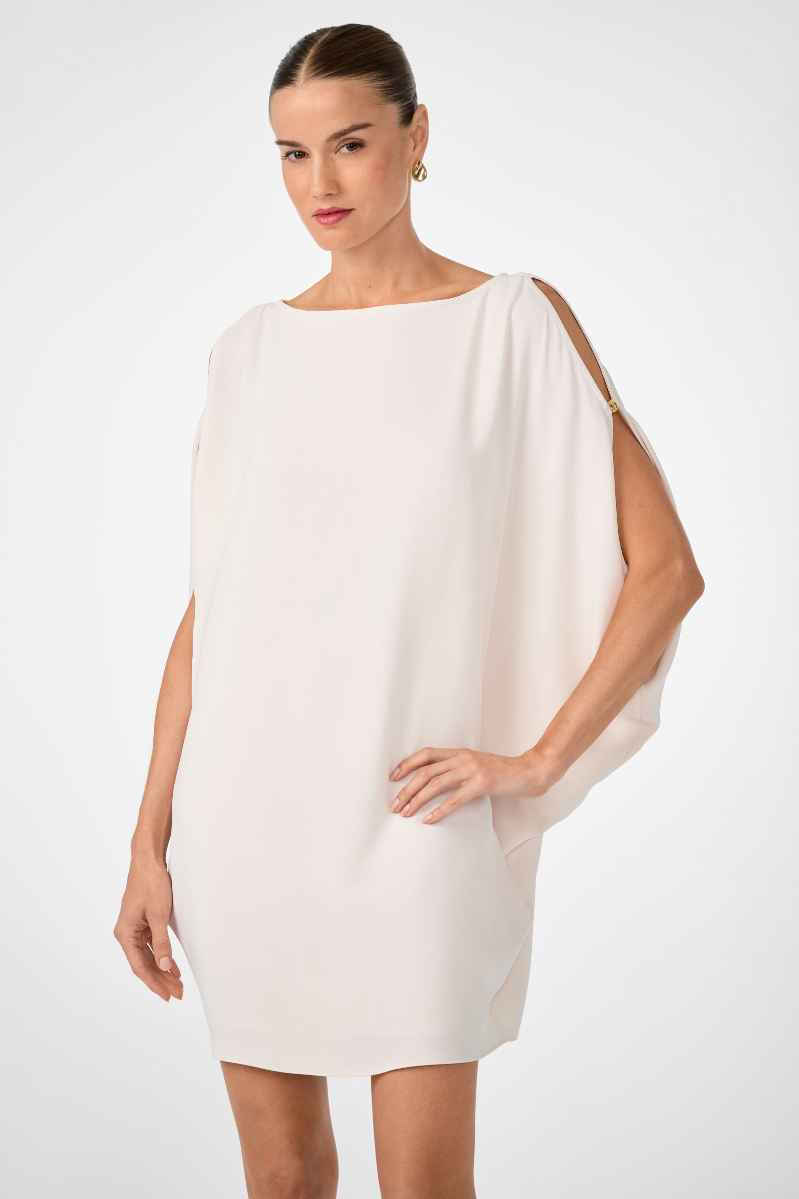 A woman in the Razu Short Split Sleeve Mini Dress, a loose short white dress with open shoulder details, and gold hoop earrings stands confidently against a plain light background.