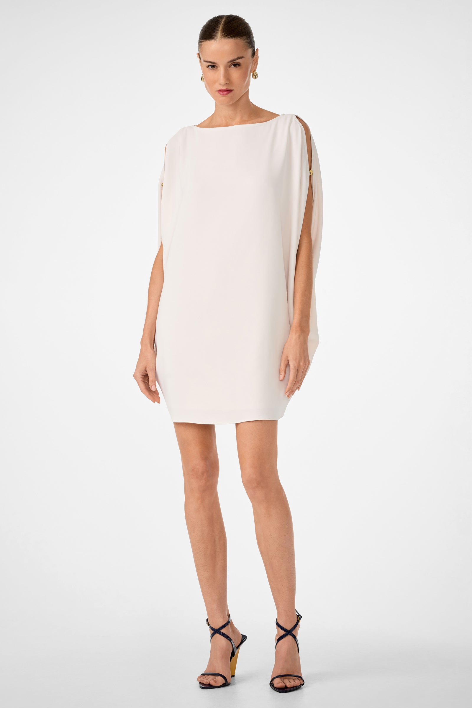 A woman stands against a white background wearing the Razu Short Split Sleeve Mini Dress in Sancerre, featuring open sleeves and gold button details at the shoulders, paired with black high-heeled sandals.
