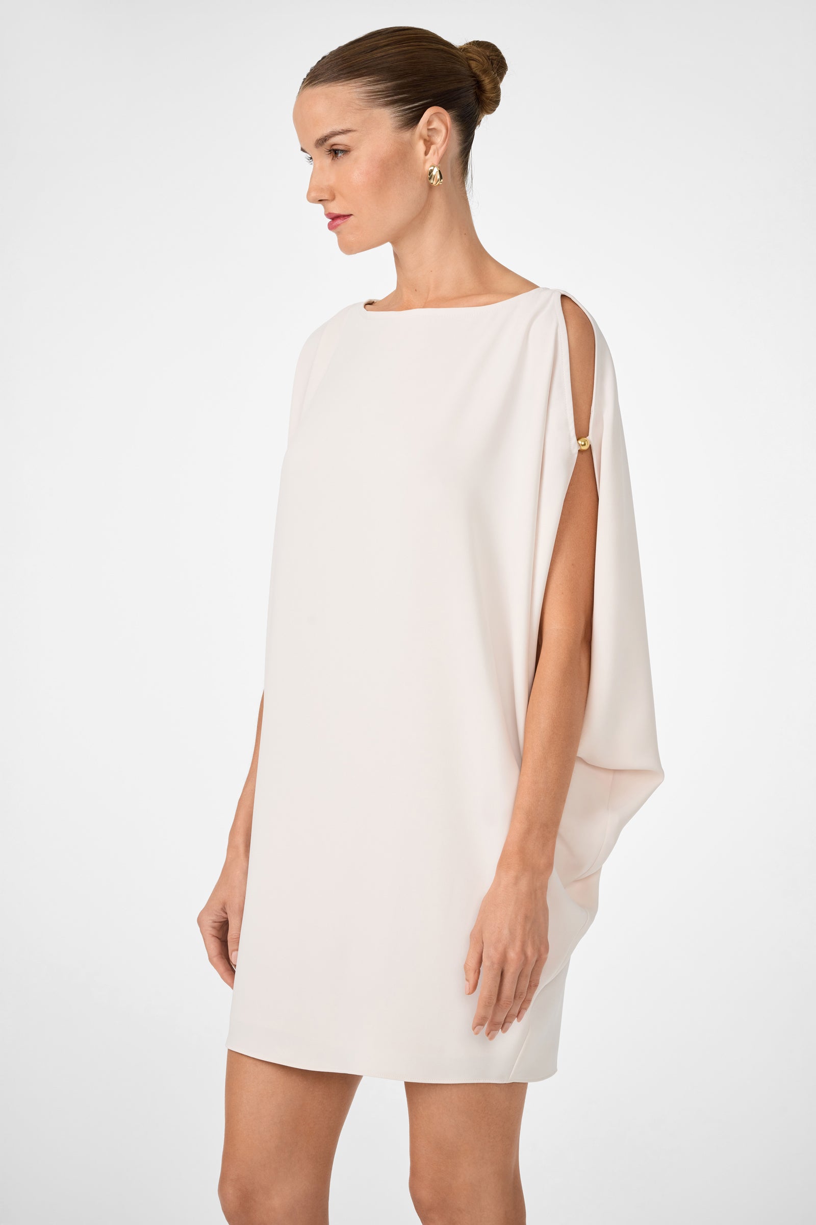 A woman wearing the Razu Short Split Sleeve Mini Dress in cream, featuring a draped design, mini length, and gold clasp detail on one shoulder. Her hair is styled in a neat bun as she stands against a plain white background.