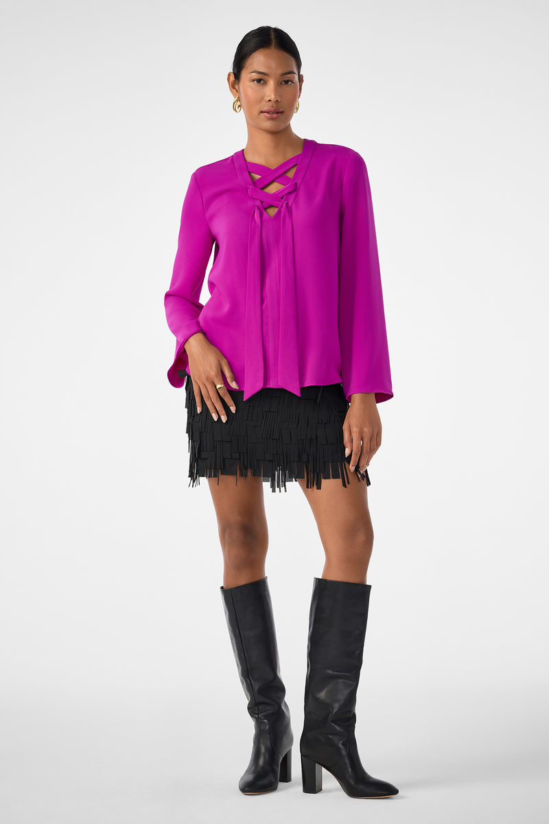 A woman poses against a plain background wearing the Rico Skirt—a black fringe mini skirt—paired with a bright magenta long-sleeve blouse with crisscross neckline detail, knee-high black boots, and gold hoop earrings.