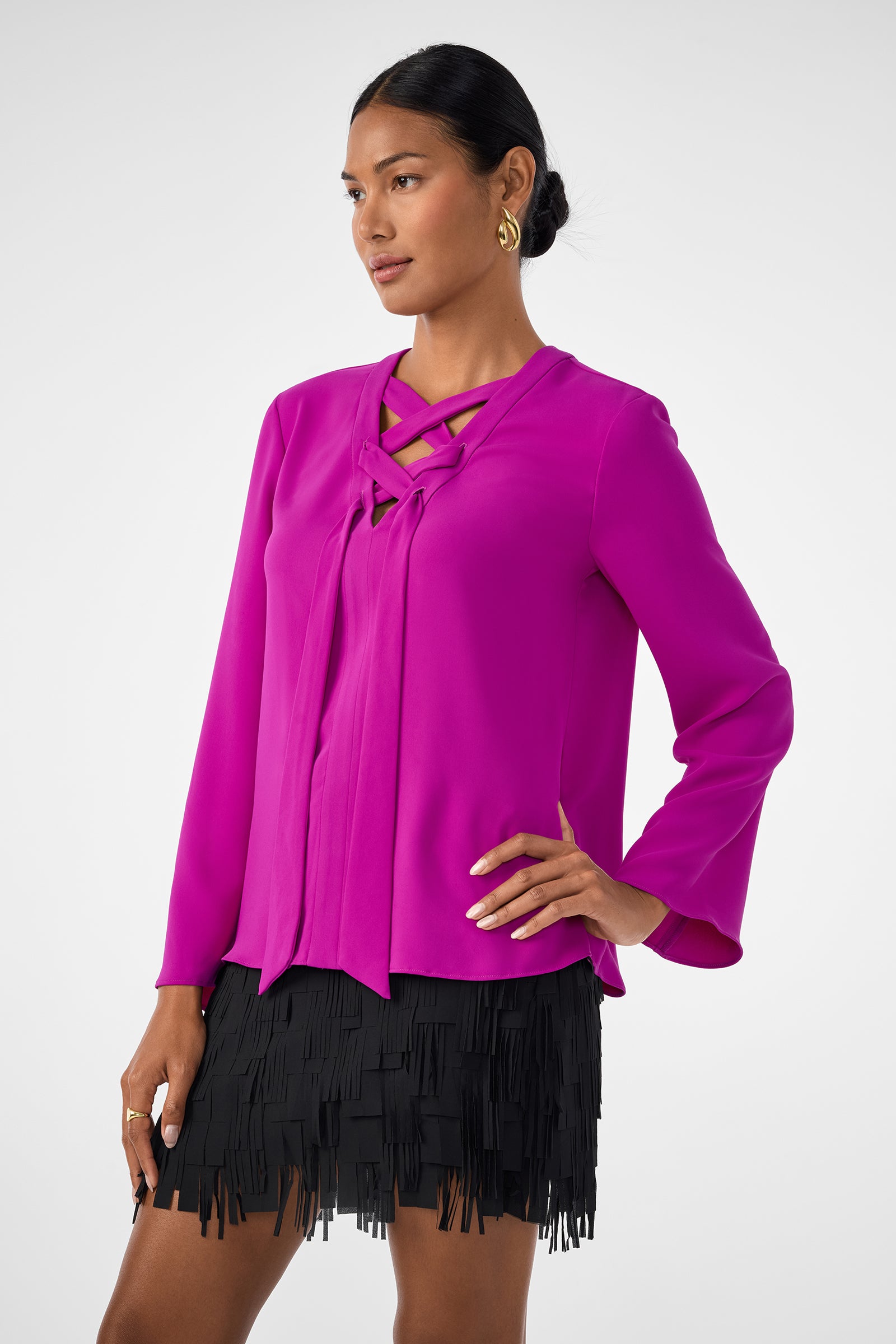 A woman stands with one hand on her hip against a light background, wearing the Picabella Top—a bright fuchsia long-sleeve blouse with crisscross neckline detail—paired with a black fringe mini skirt.