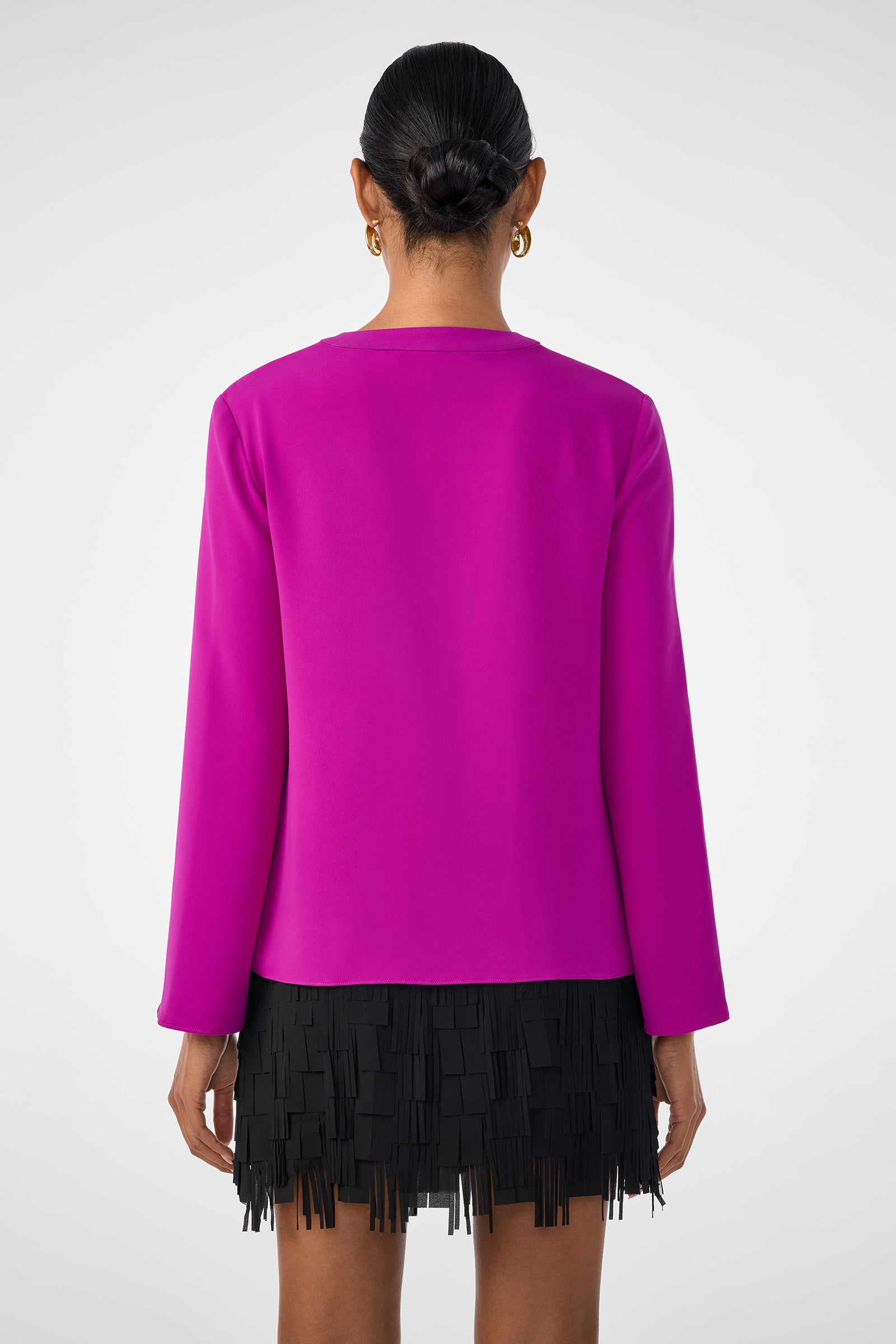 A person with dark hair in a sleek bun is seen from behind, wearing the magenta Picabella Top with long sleeves over a black fringed skirt, set against a plain light background.