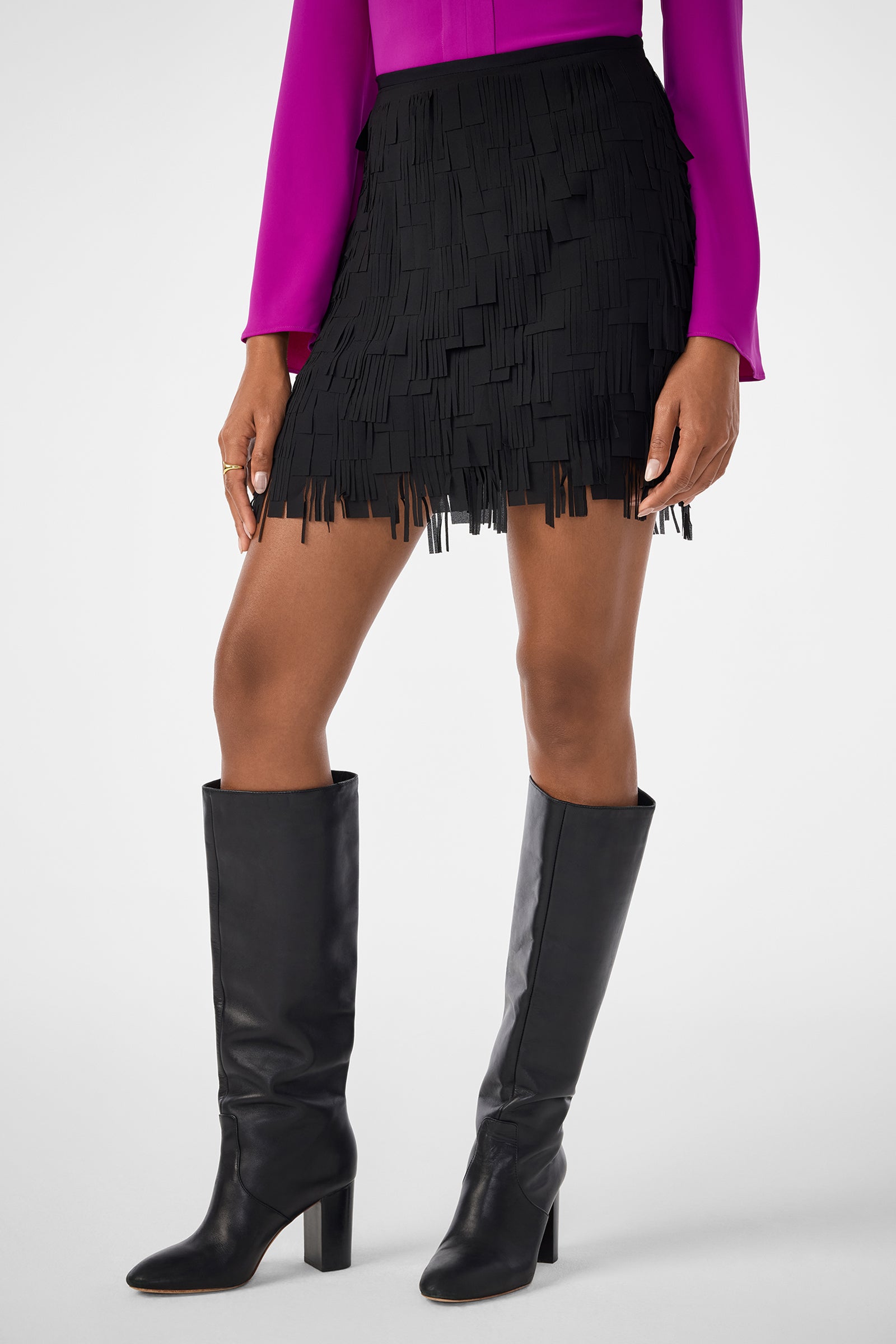 A person wearing the Rico Skirt—a black fringed mini skirt—paired with a bright pink long-sleeve top and knee-high black heeled boots, stands against a plain light background.