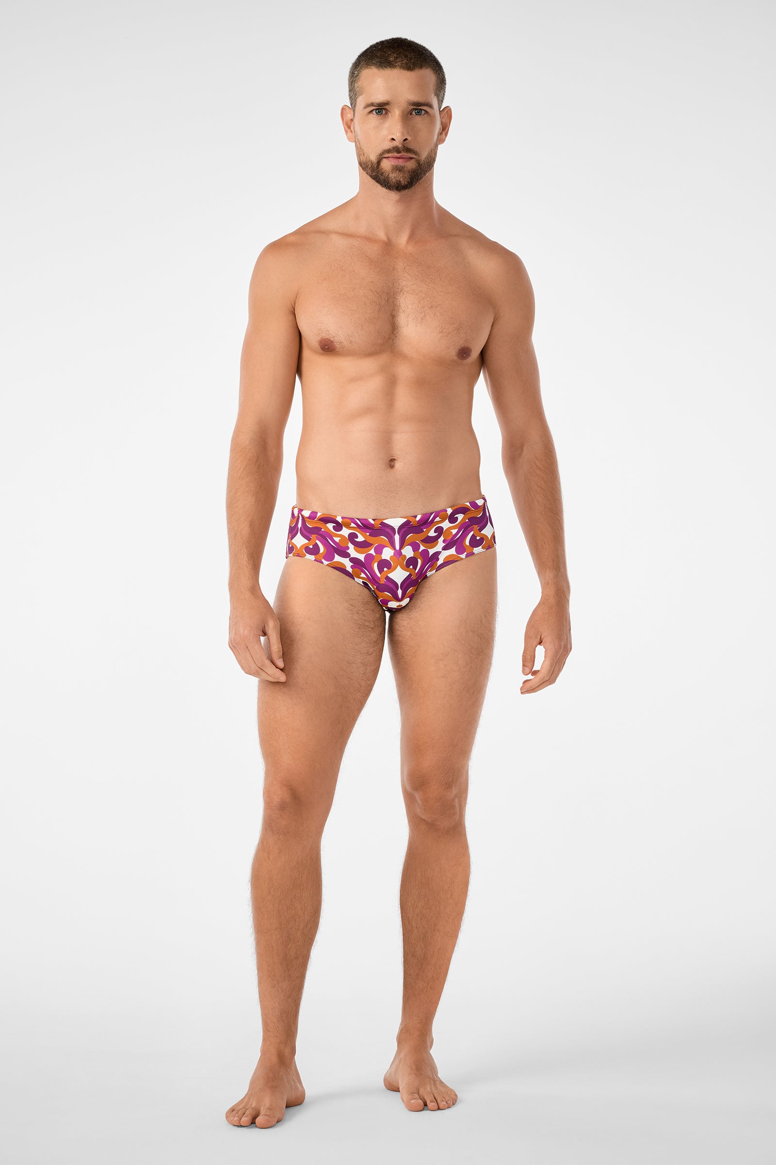 A man with short hair and a beard stands barefoot against a plain background, wearing the Bondi Swim Brief in red, purple, and yellow patterns. He is facing forward with a neutral expression and arms relaxed at his sides.