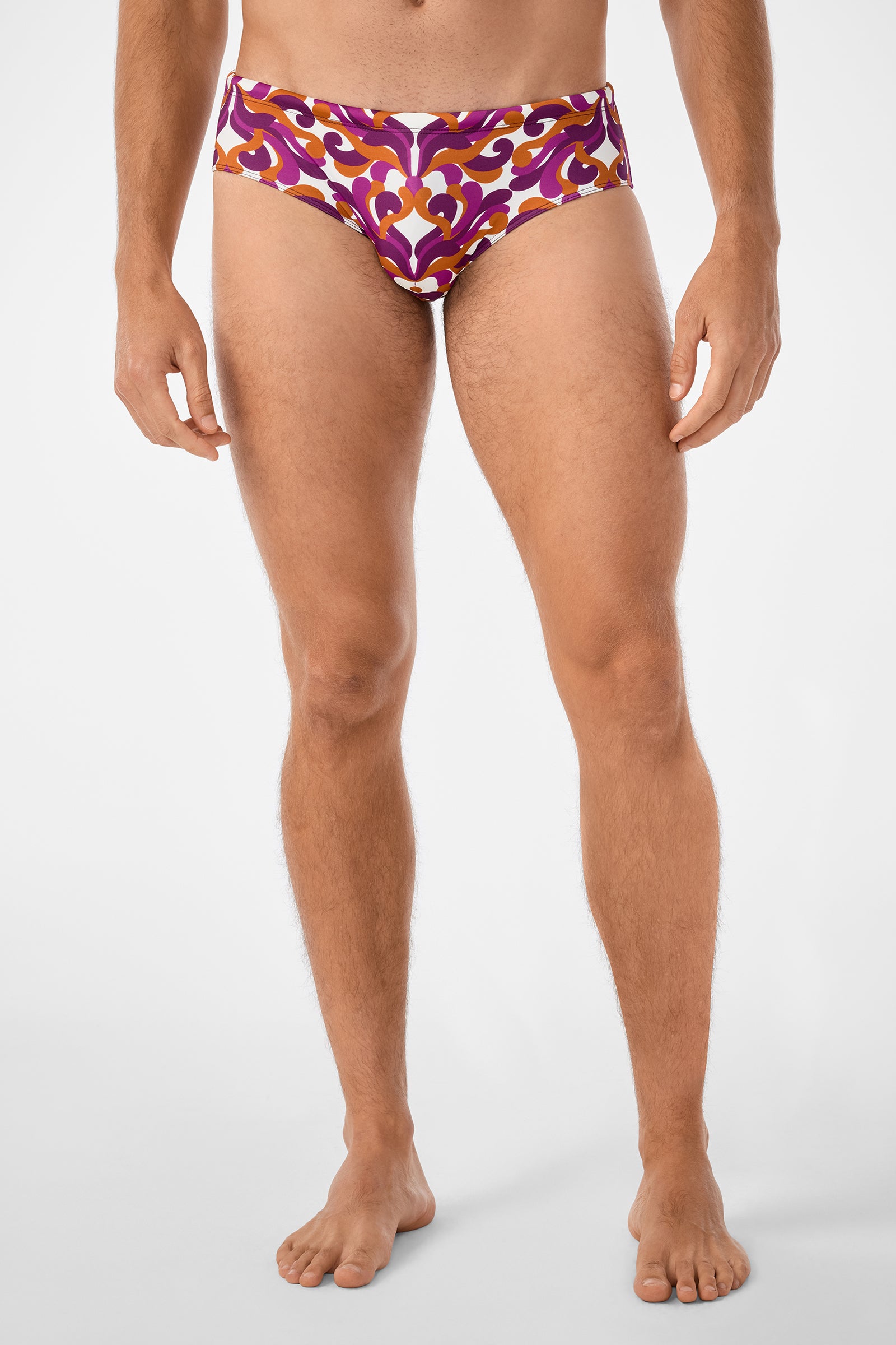 A barefoot man models the Bondi Swim Brief, featuring a retro abstract pattern in orange, purple, and white, set against a plain white background.