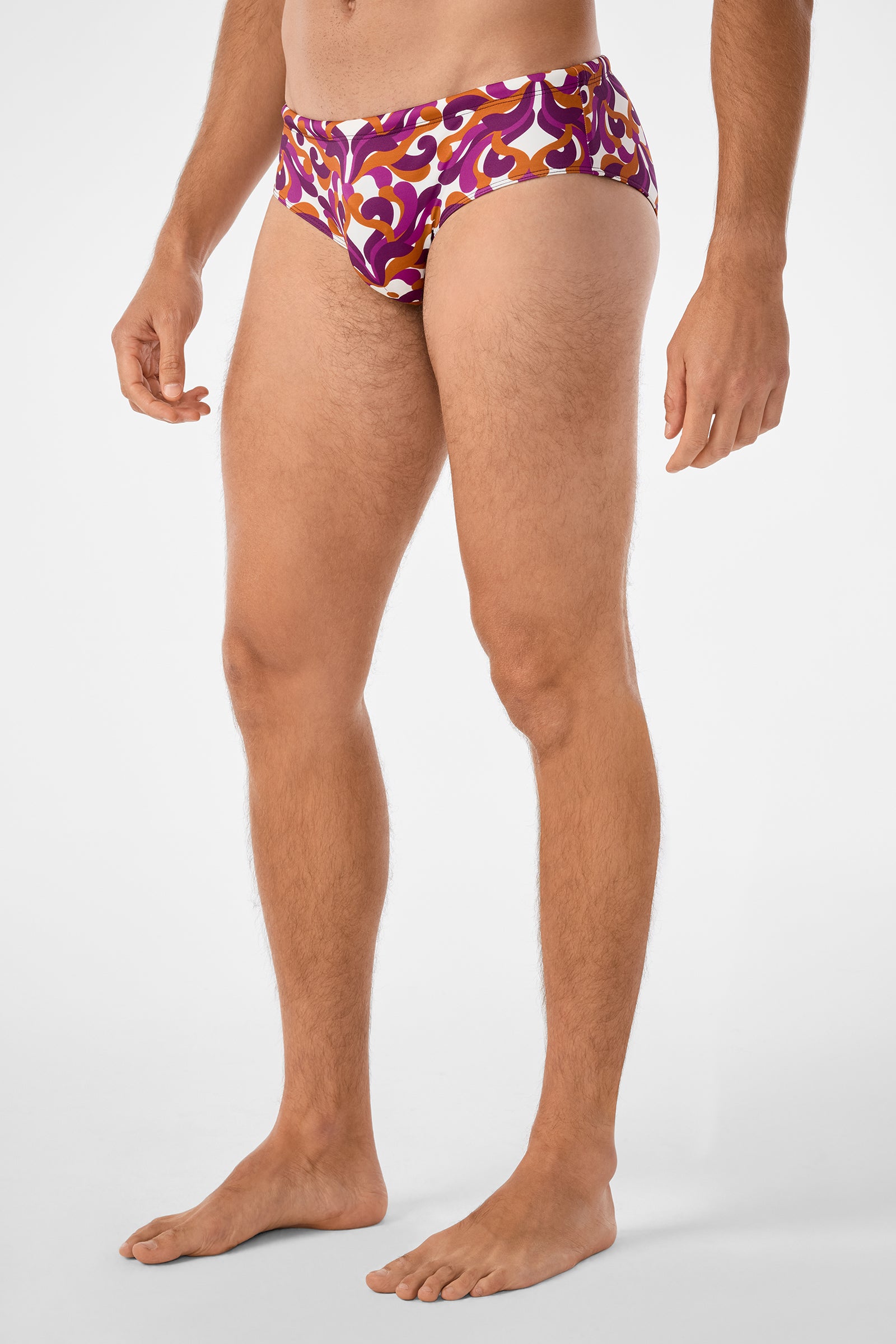 A man models the Bondi Swim Brief, featuring a bold, colorful pattern in purple, gold, and white. He stands barefoot against a plain white background, shown from upper chest to feet.