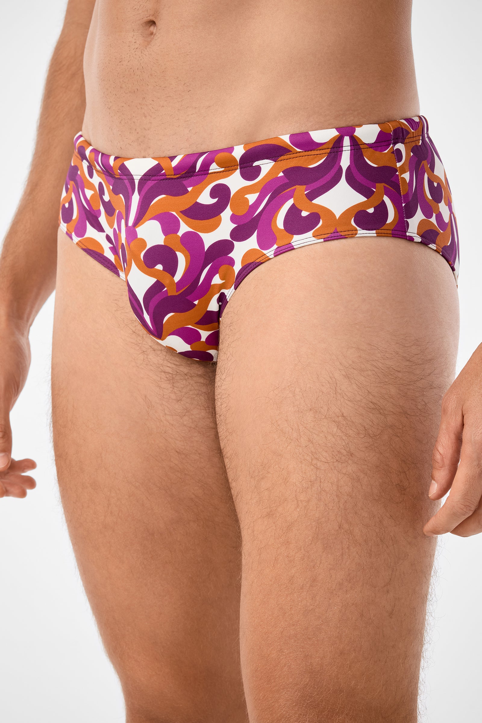 A person models the Bondi Swim Brief, featuring a bold purple, orange, and white swirling pattern. The image is cropped to display the torso and upper thighs against a plain background.