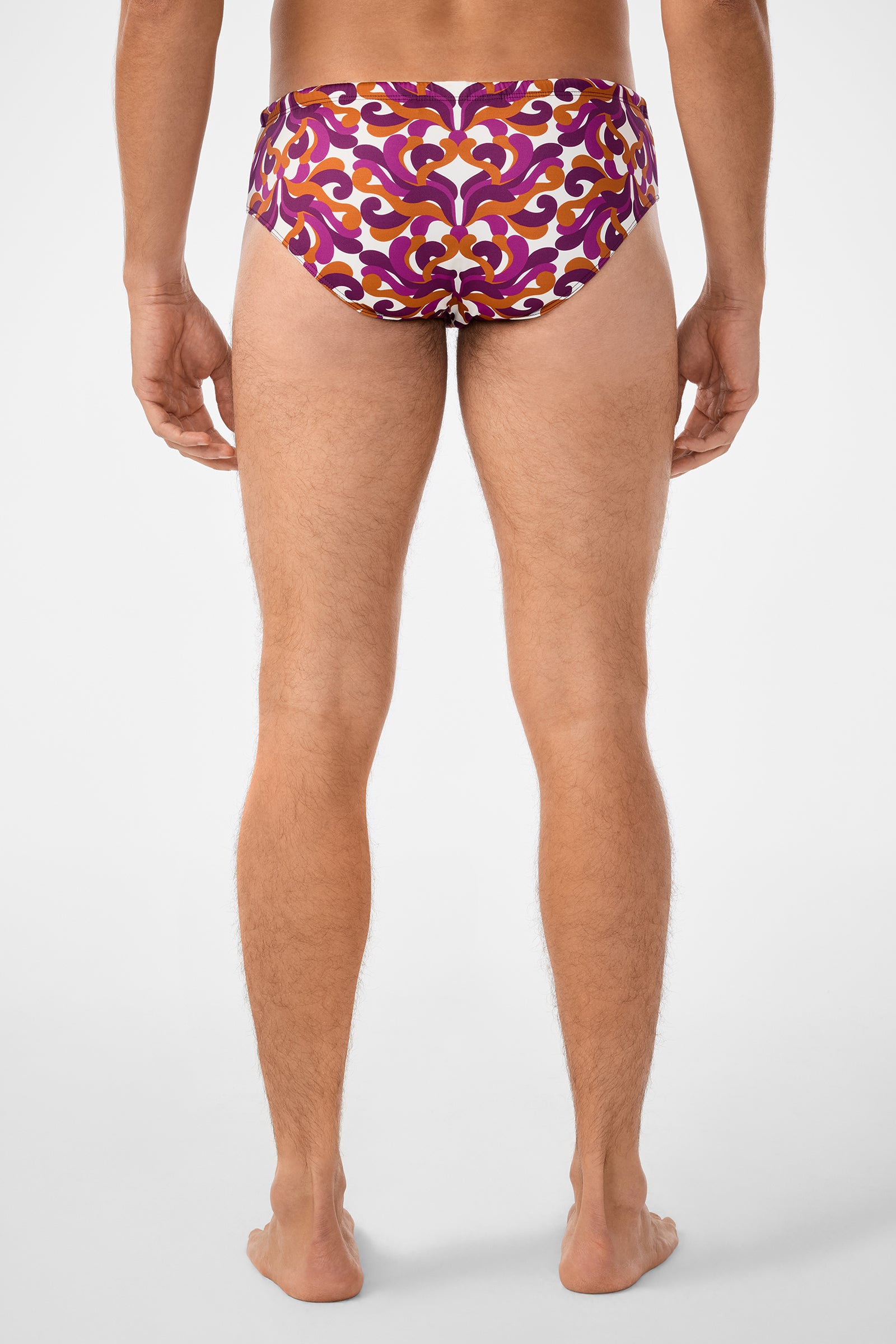 A man stands barefoot with his back to the camera, wearing the Bondi Swim Brief featuring a colorful pattern in purple, orange, and white against a plain white background.