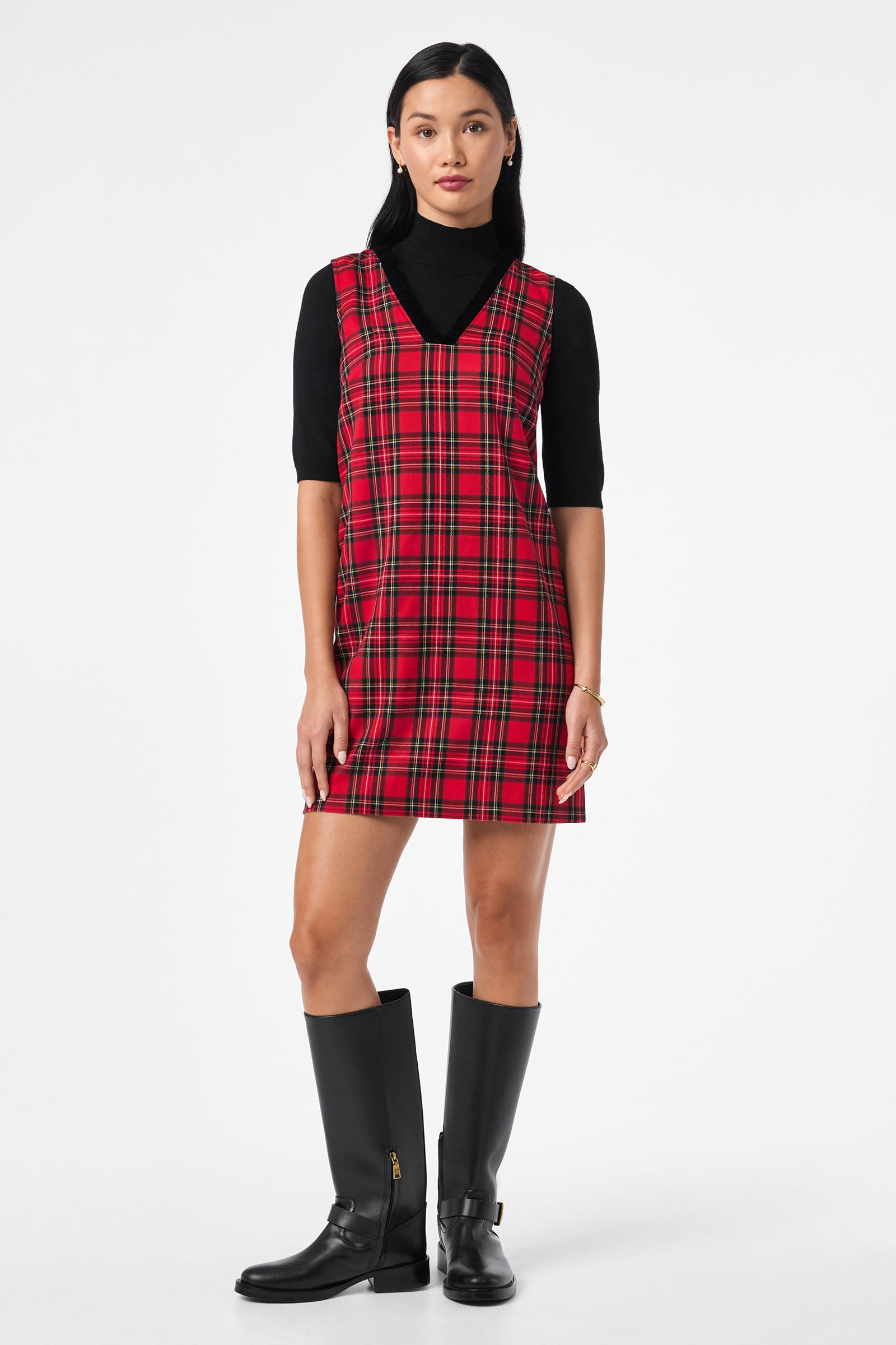 A woman stands facing forward in the Revah Dress, a sleeveless red plaid style layered over a black turtleneck and paired with knee-high black boots. She has straight dark hair and minimal jewelry, posing against a plain white background.