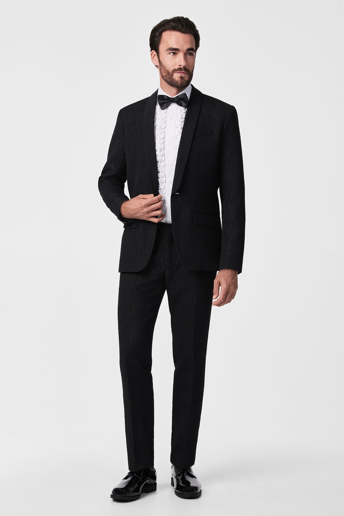 A man with short dark hair stands against a white background wearing the Thurston Blazer, a black textured tuxedo, paired with a ruffled white dress shirt, black bow tie, and black dress shoes. He gazes slightly to the side.