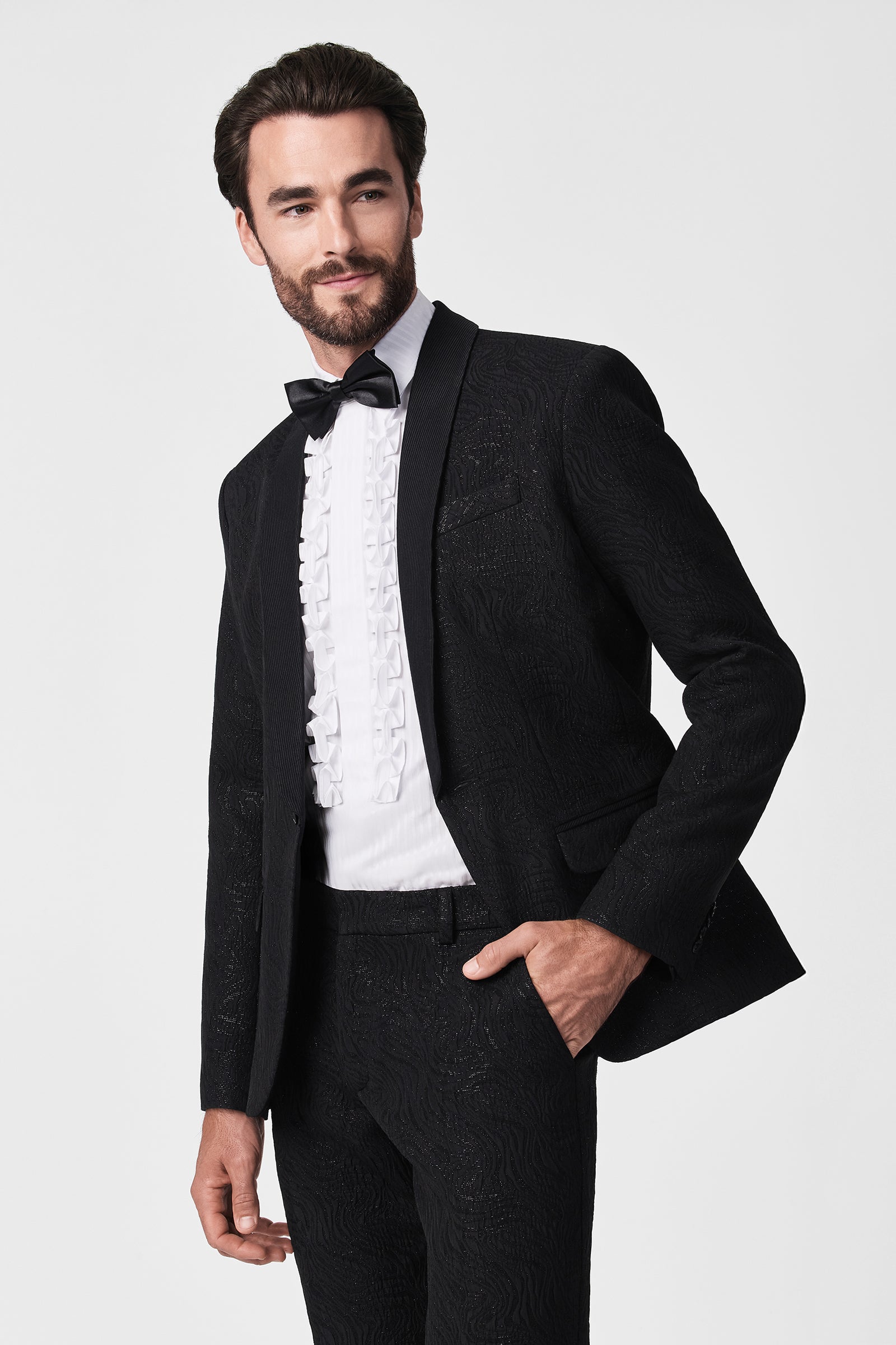 A man models the Adrian Tux Trouser, paired with a textured black tuxedo jacket, black bow tie, and white ruffle dress shirt, posing with one hand in his pocket against a plain light background.