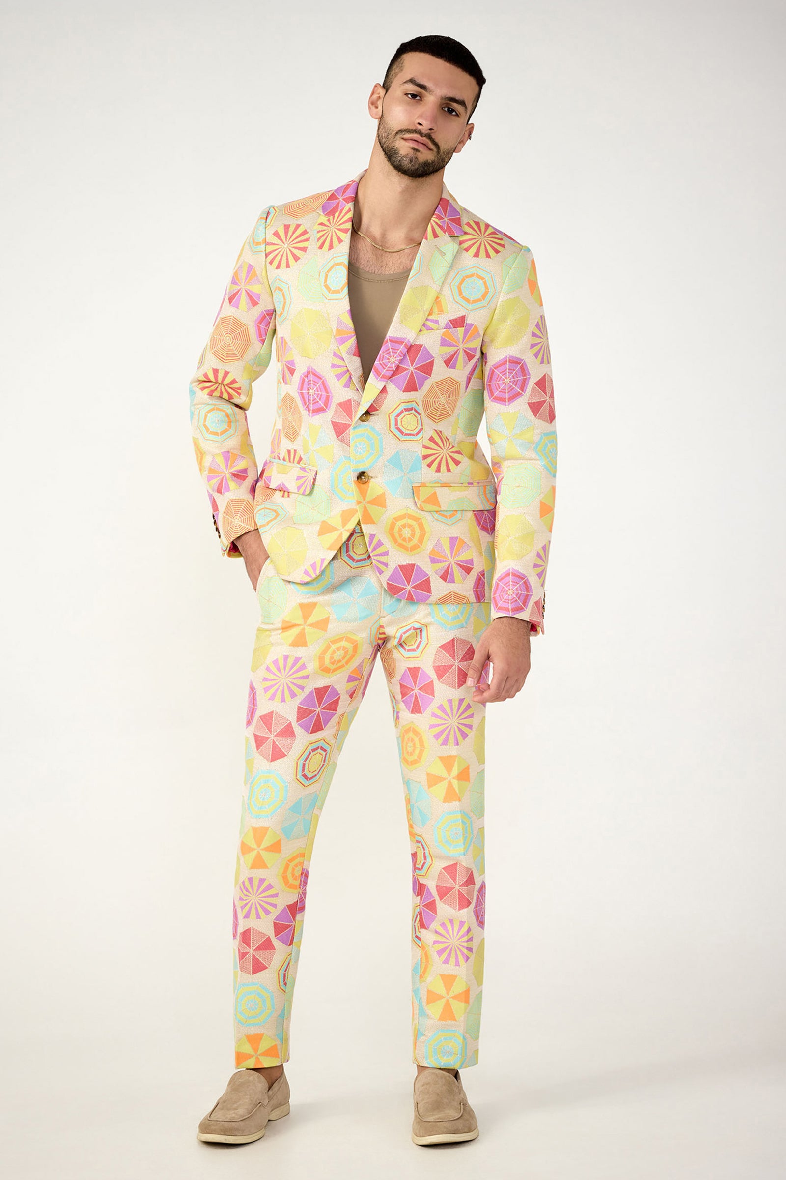 A man wears the Thurston Blazer - Parapluie Jacquard, featuring bold pastel geometric star and swirl patterns, paired with a beige shirt and suede shoes, set against a plain light background.