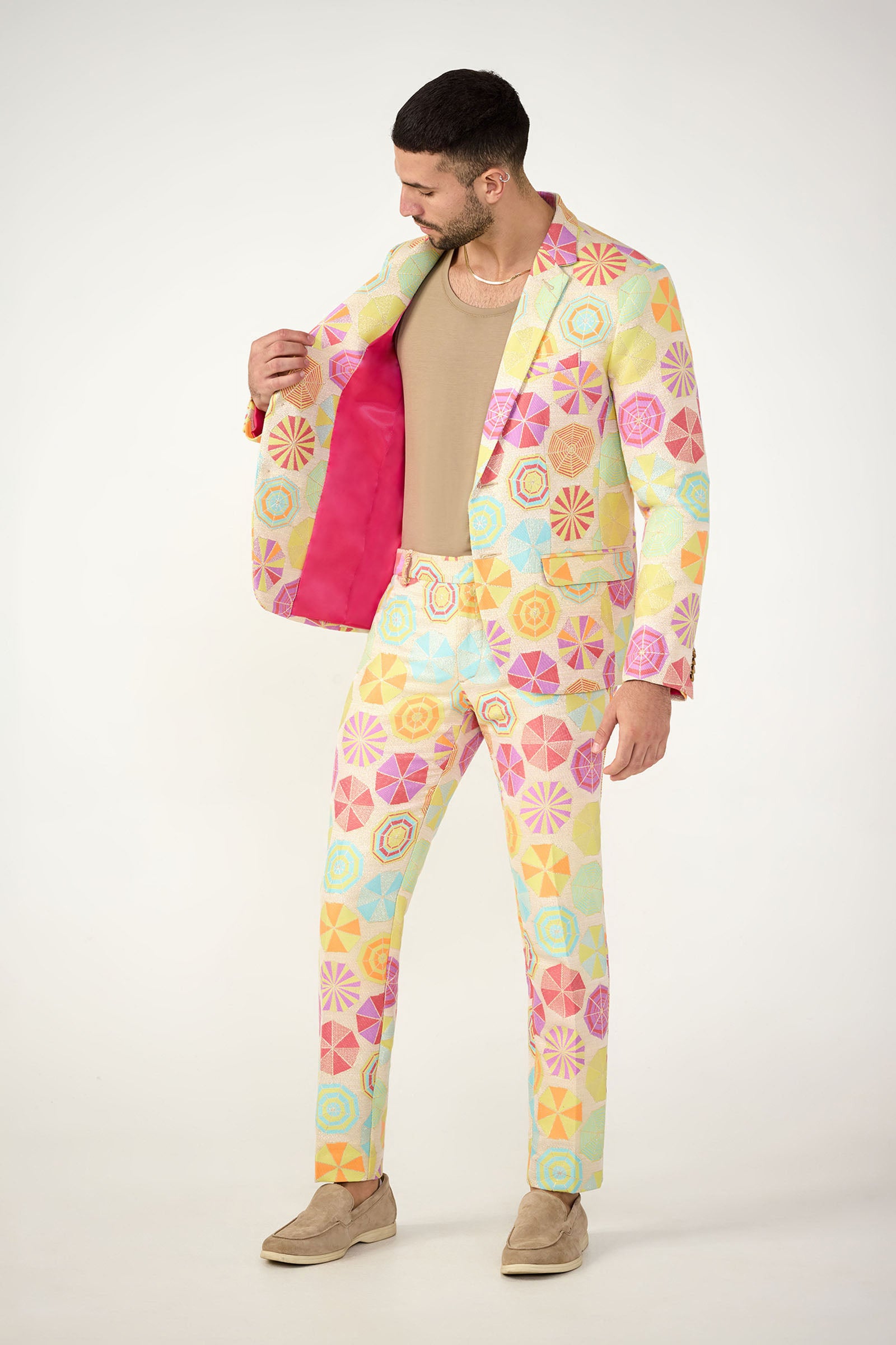 A man models the Thurston Blazer - Parapluie Jacquard, featuring playful pastel geometric patterns and a bright pink lining, paired with a beige shirt, tan loafers, and gold chain against a white background.