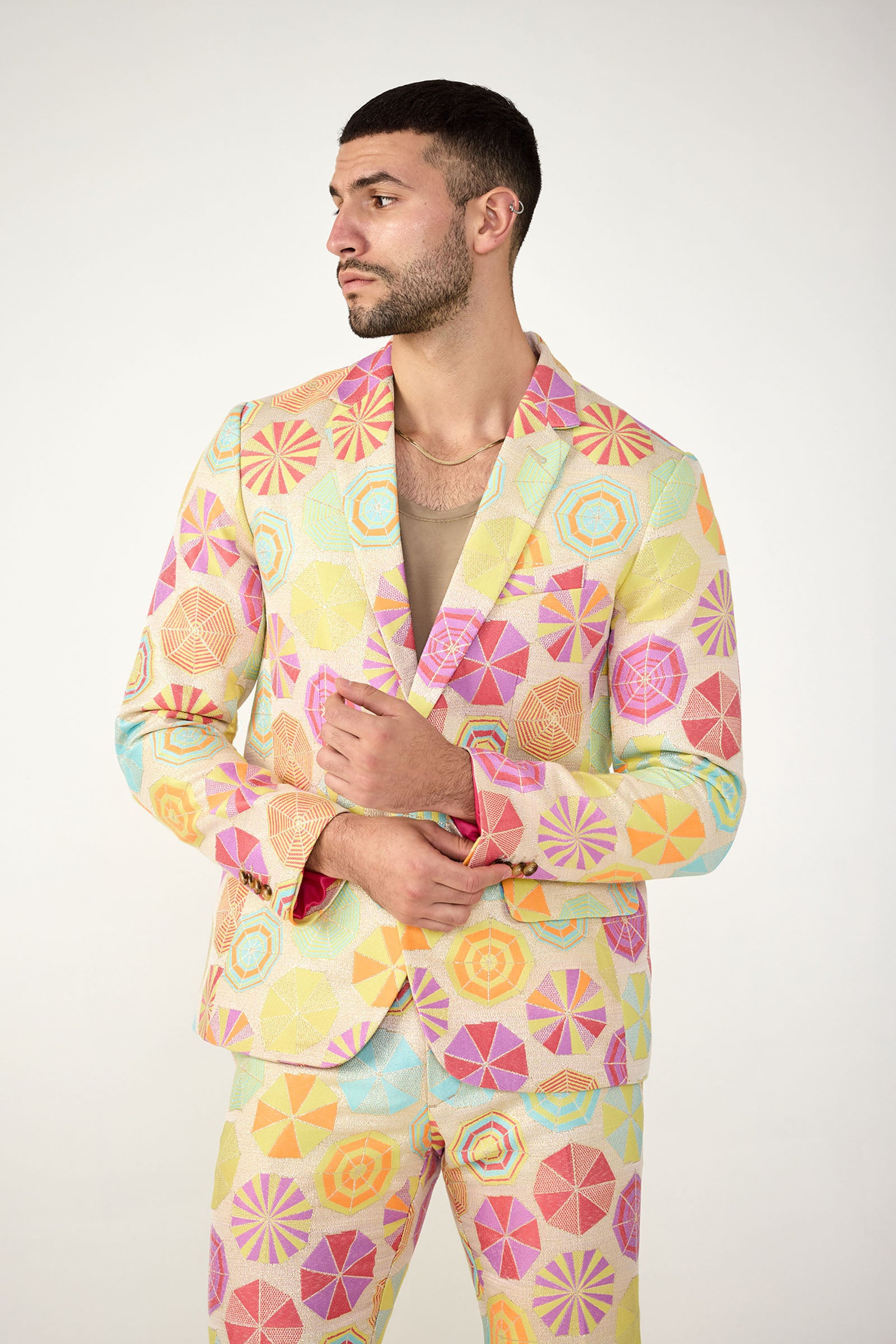A man wears the Thurston Blazer in Parapluie Jacquard, featuring colorful geometric pastel patterns, as he stands against a plain background and looks left while adjusting his jacket.