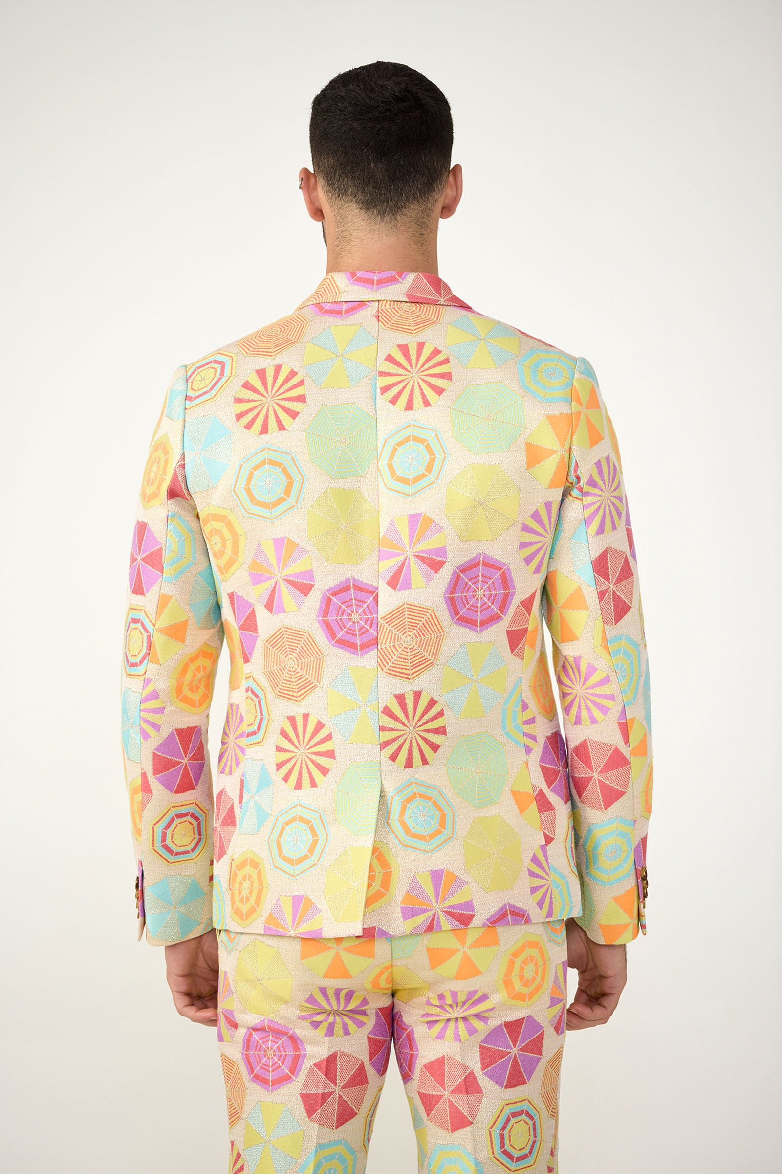 A person stands facing away, wearing the Thurston Blazer - Parapluie Jacquard, a colorful suit featuring a geometric pattern of circles and polygons in pastel pink, yellow, blue, and orange against a plain white background.