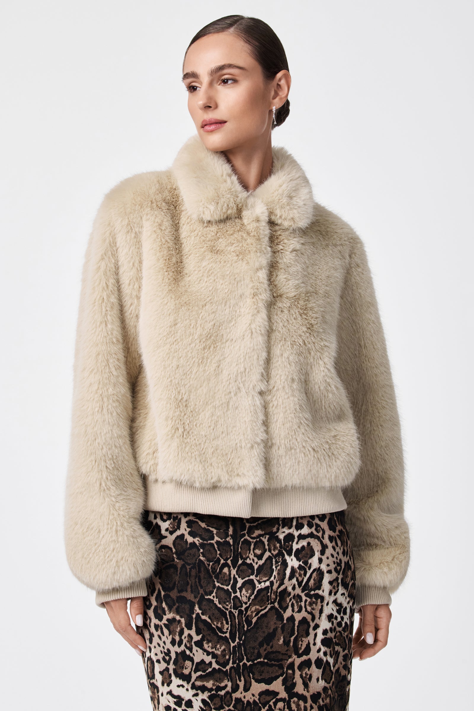 A woman wears the Morris Short Faux Fur Coat, a cream-colored jacket with a high collar, paired with a leopard print skirt, against a plain white background.