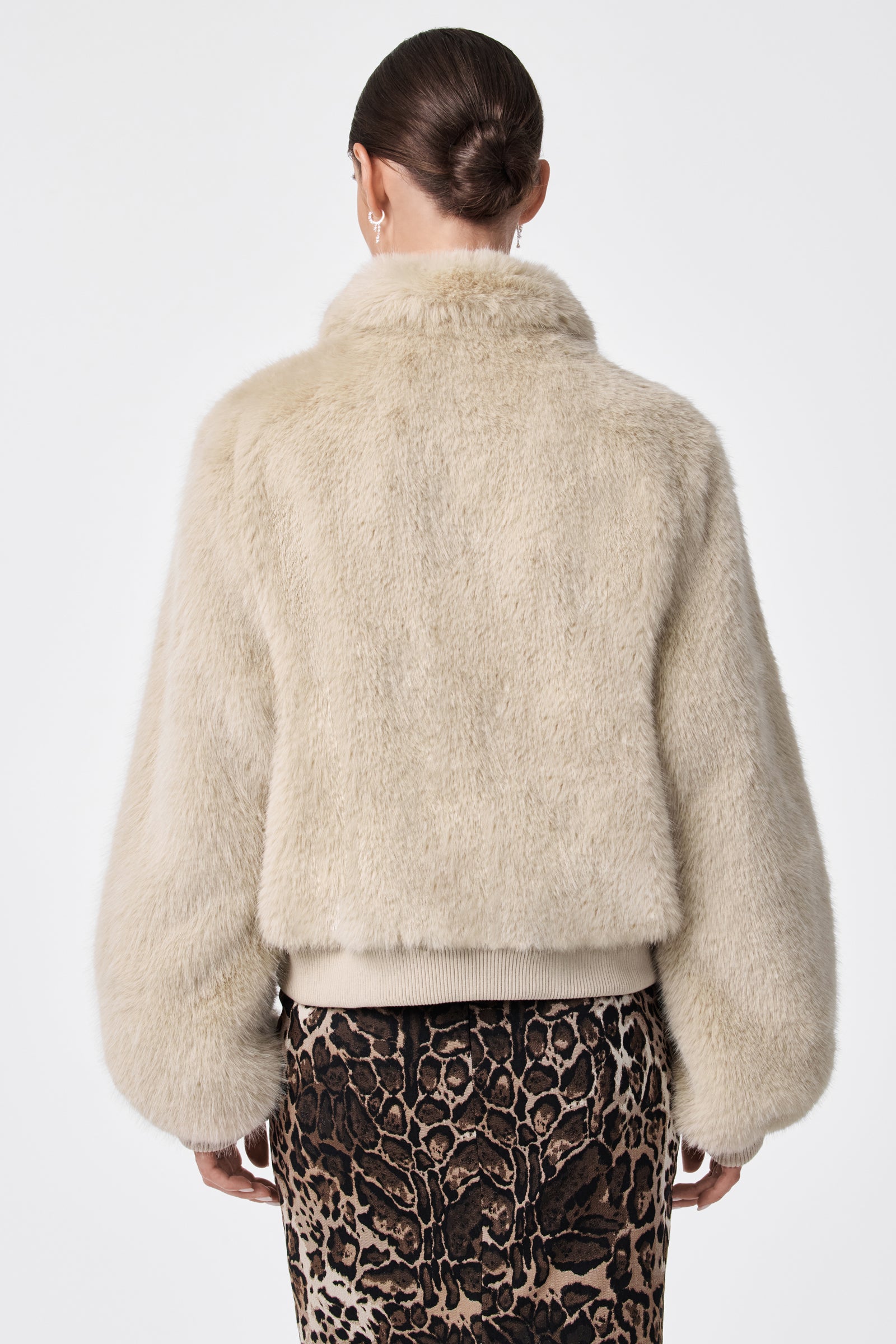 A person with dark hair in a low bun models the Morris Short Faux Fur Coat in light beige and a brown leopard print skirt, seen from the back against a plain white background.