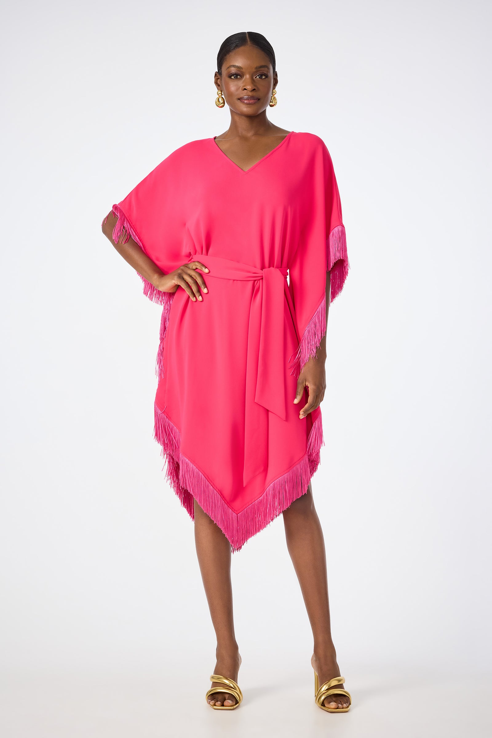 A woman poses against a white background wearing the Toulouse Short-Sleeve Fringe Dress in Cannes Coral, featuring fringed edges, a V-neckline, and matching waist tie. She completes the look with gold hoop earrings and gold heeled sandals.