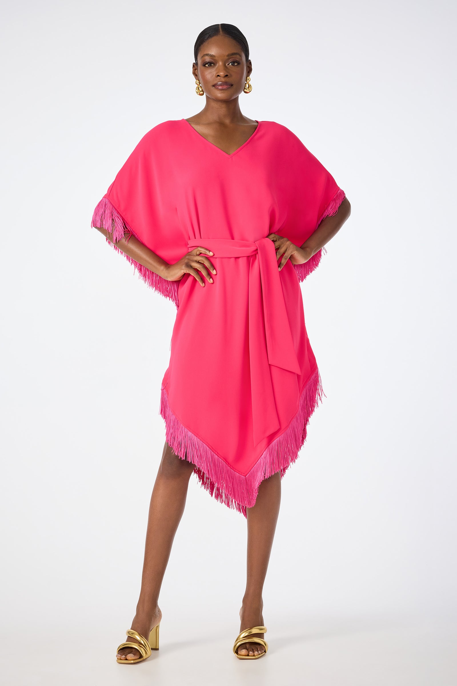 A woman stands confidently in the Toulouse Short-Sleeve Fringe Dress—a bright pink V-neck style with a matching belt—paired with gold heeled sandals and hoop earrings, posing against a plain white background.