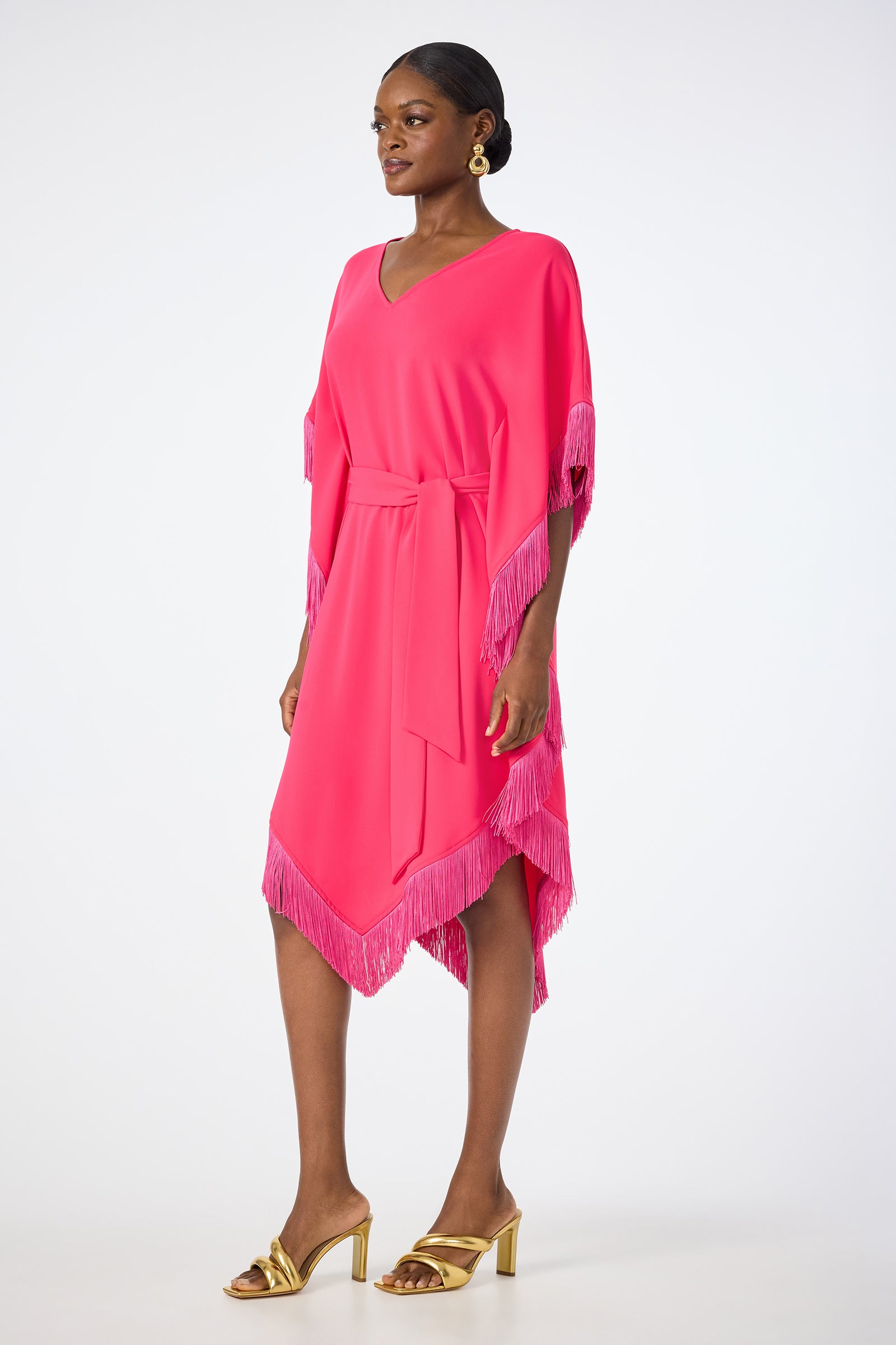 A woman wears the Toulouse Short-Sleeve Fringe Dress in bright pink, featuring a V-neck, knee-length cut, fringed sleeves and hem, and a matching belt. She pairs it with gold heeled sandals against a plain white background.