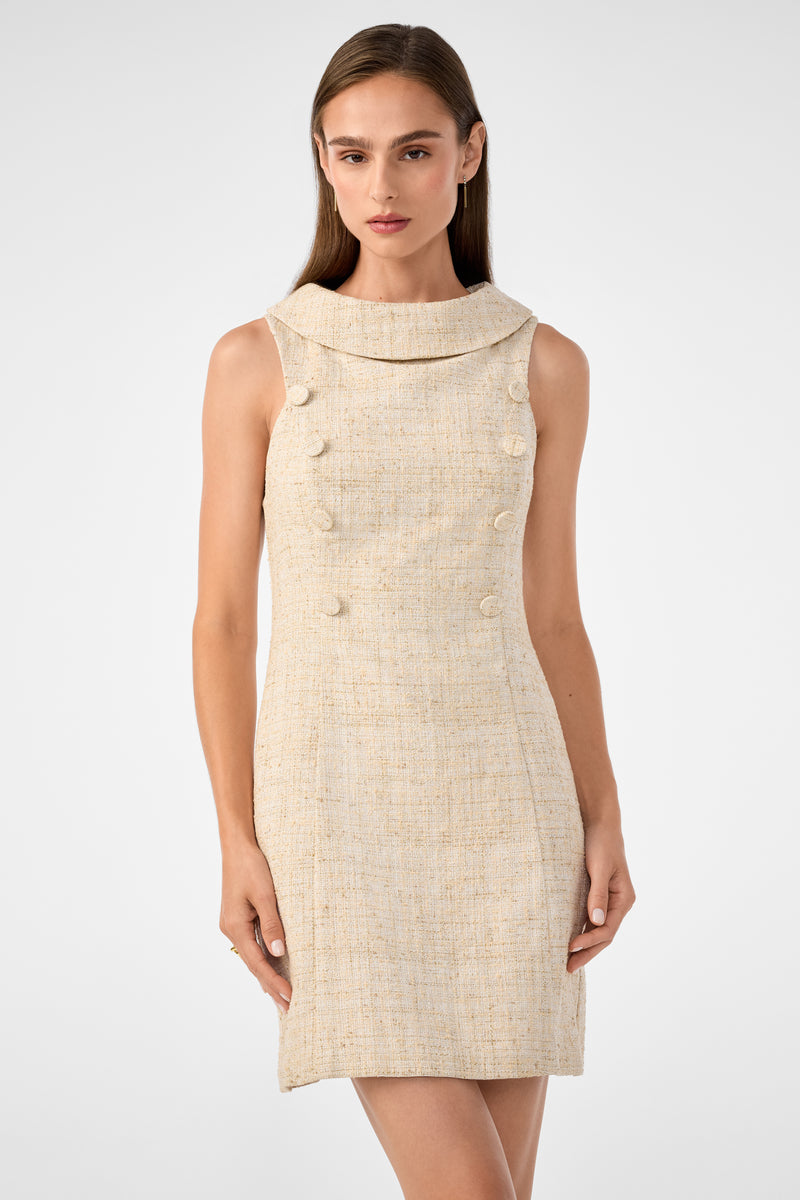 A woman with straight brown hair models the Beziers Dress, a sleeveless beige tweed style with a wide collar and front decorative buttons, standing against a plain light gray background.