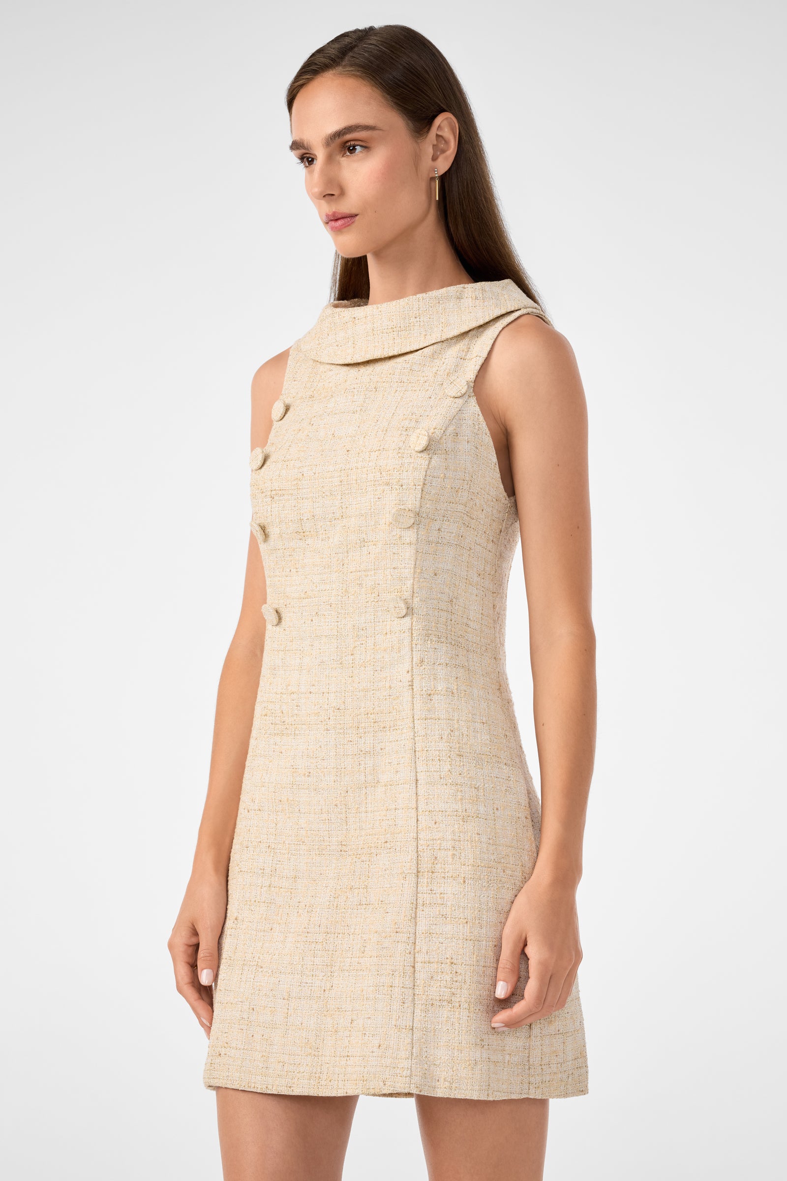 A woman wears the Beziers Dress—a sleeveless beige textured mini dress featuring a wide, structured collar and decorative front buttons—while standing against a plain light background.