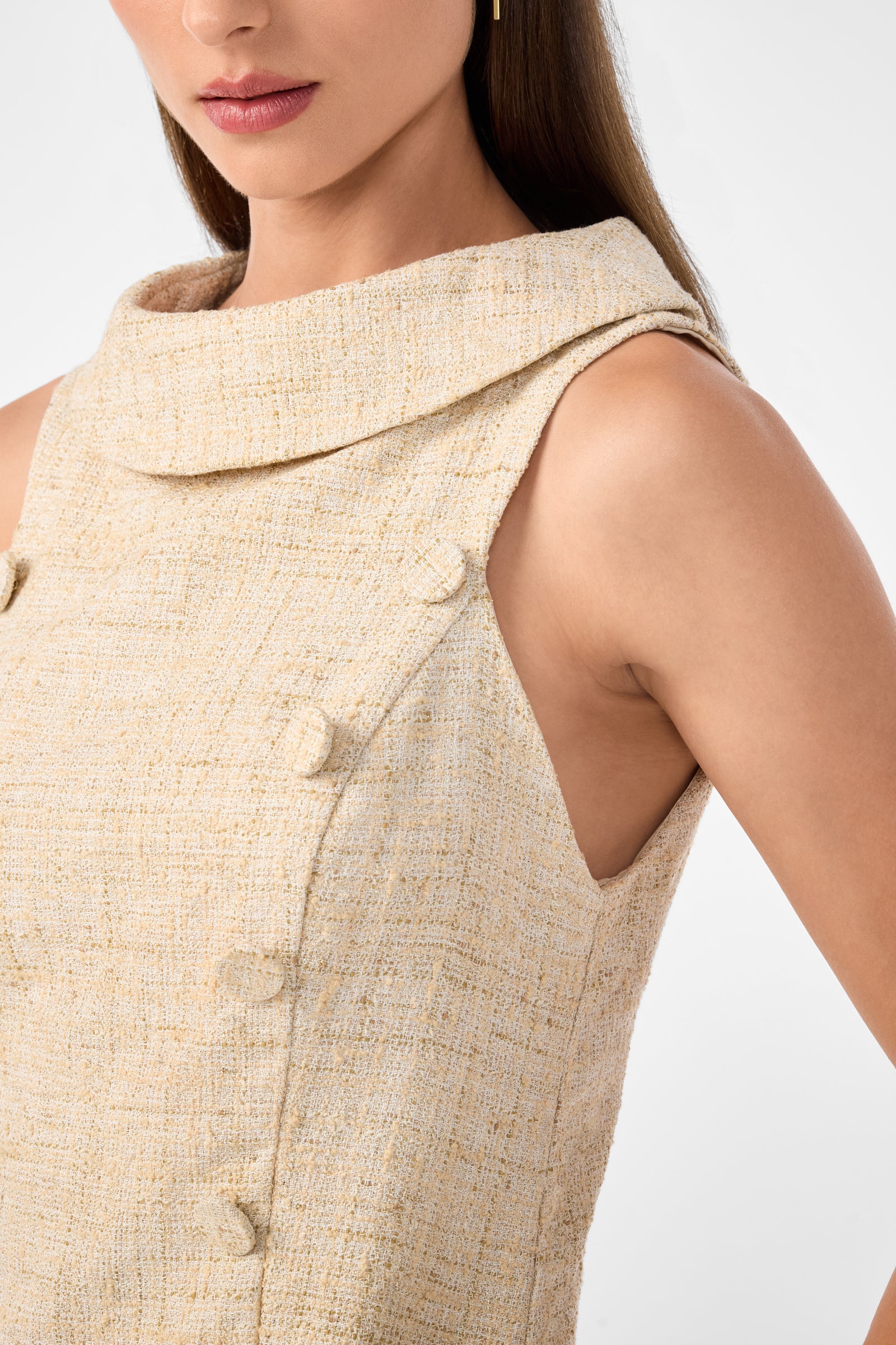 A close-up of a woman wearing the Beziers Dress, a sleeveless beige textured piece with a wide collar and decorative buttons, shown from shoulders to mid-torso against a plain background.