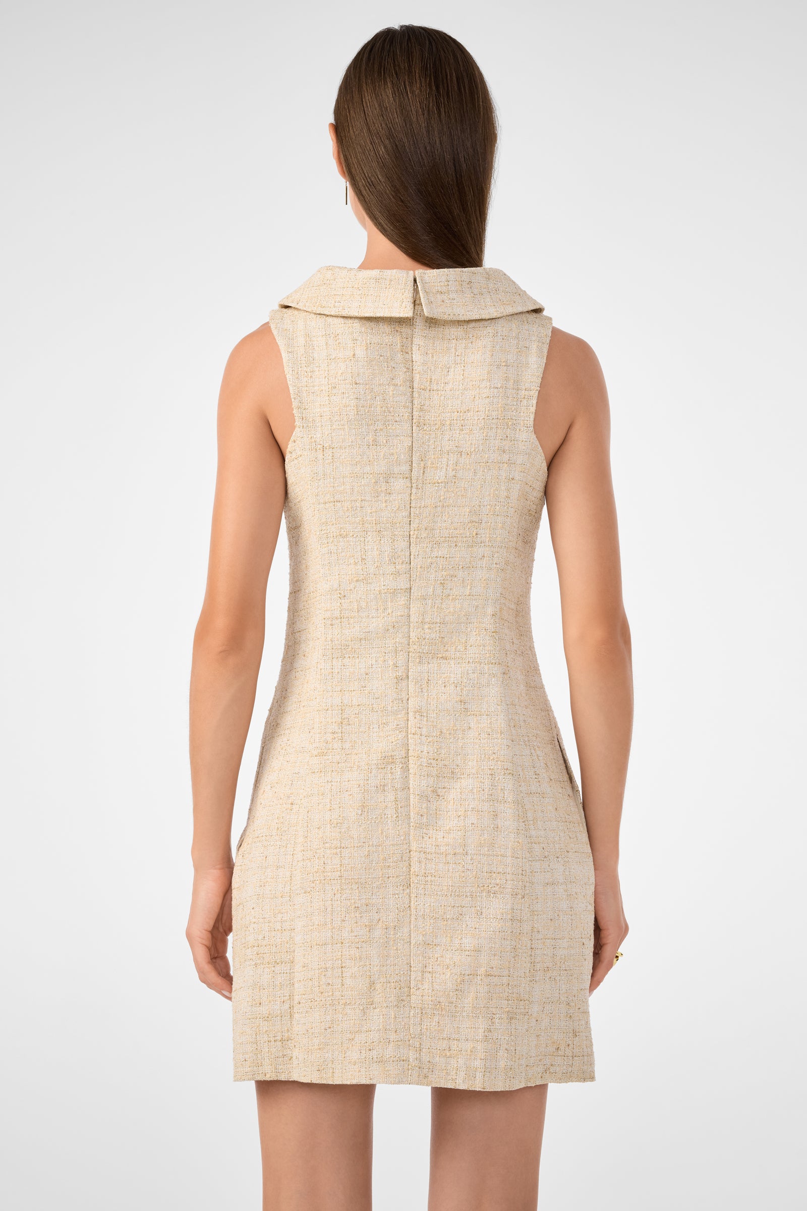 A woman with straight brown hair stands facing away, wearing the Beziers Dress—a sleeveless, fitted beige textured dress with a wide collar that falls above the knees—against a plain light background.