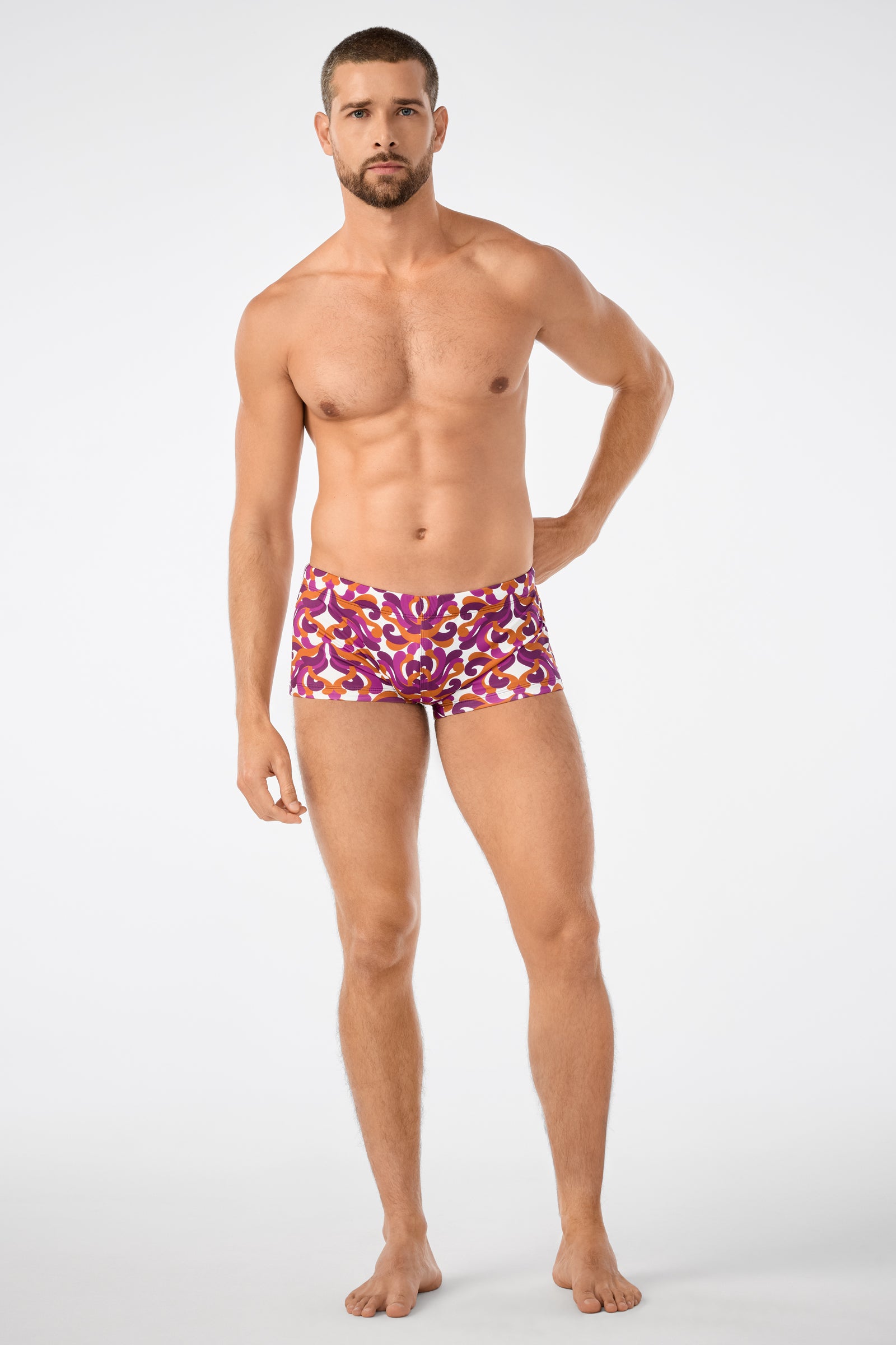 A man with short hair and a beard stands barefoot against a plain background, wearing Jet Set Swim Trunk in colorful pink, red, and yellow patterns.