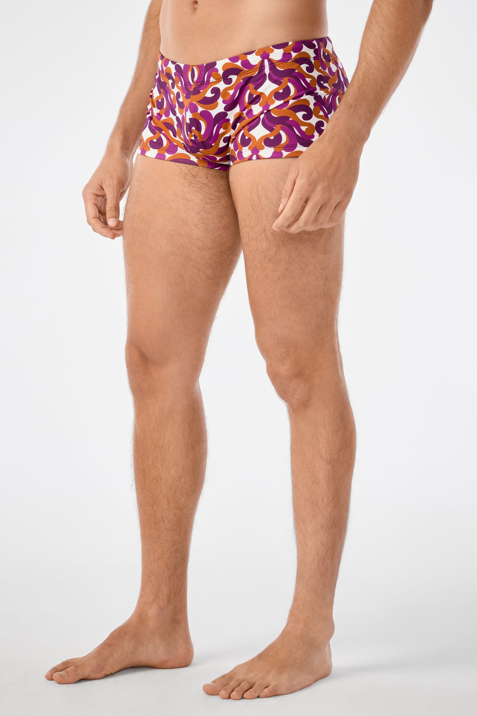 A man stands barefoot against a plain white background wearing Jet Set Swim Trunk, which features a colorful, fitted design with a bold purple, red, and yellow abstract pattern. His face is not visible.