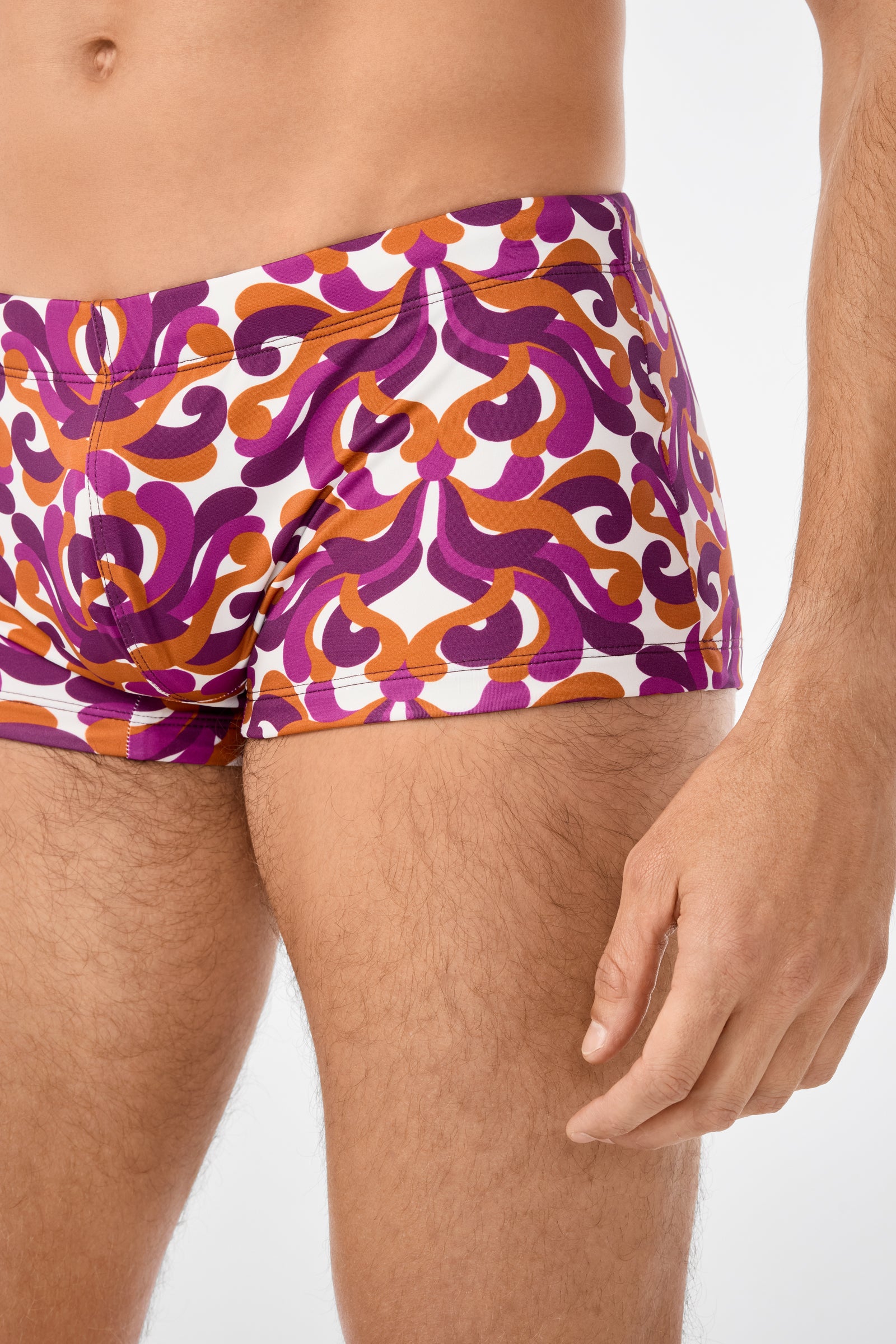 A person modeling Jet Set Swim Trunk, featuring a vibrant purple, orange, and white abstract pattern in a retro style, shown from mid-chest to mid-thigh against a plain background.