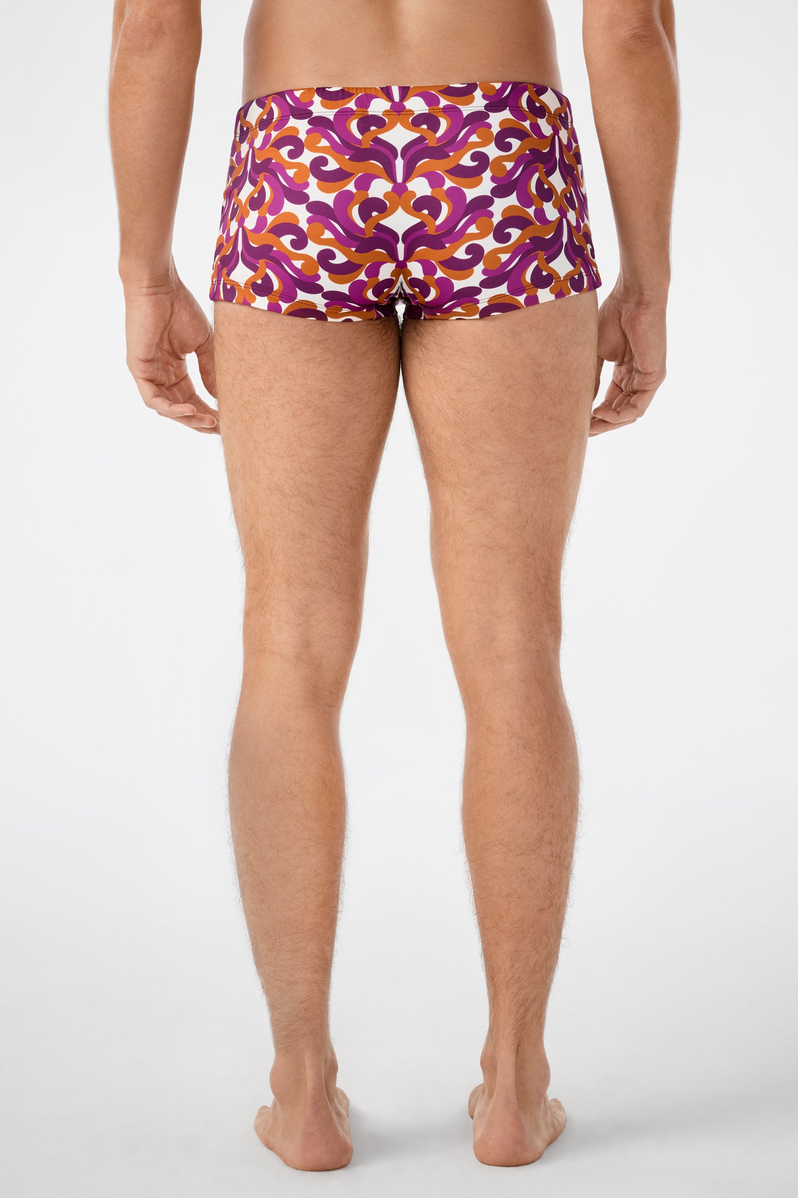 A person faces away from the camera wearing Jet Set Swim Trunk, featuring a bold maroon, orange, and white abstract swirl pattern. The background is plain and light-colored.