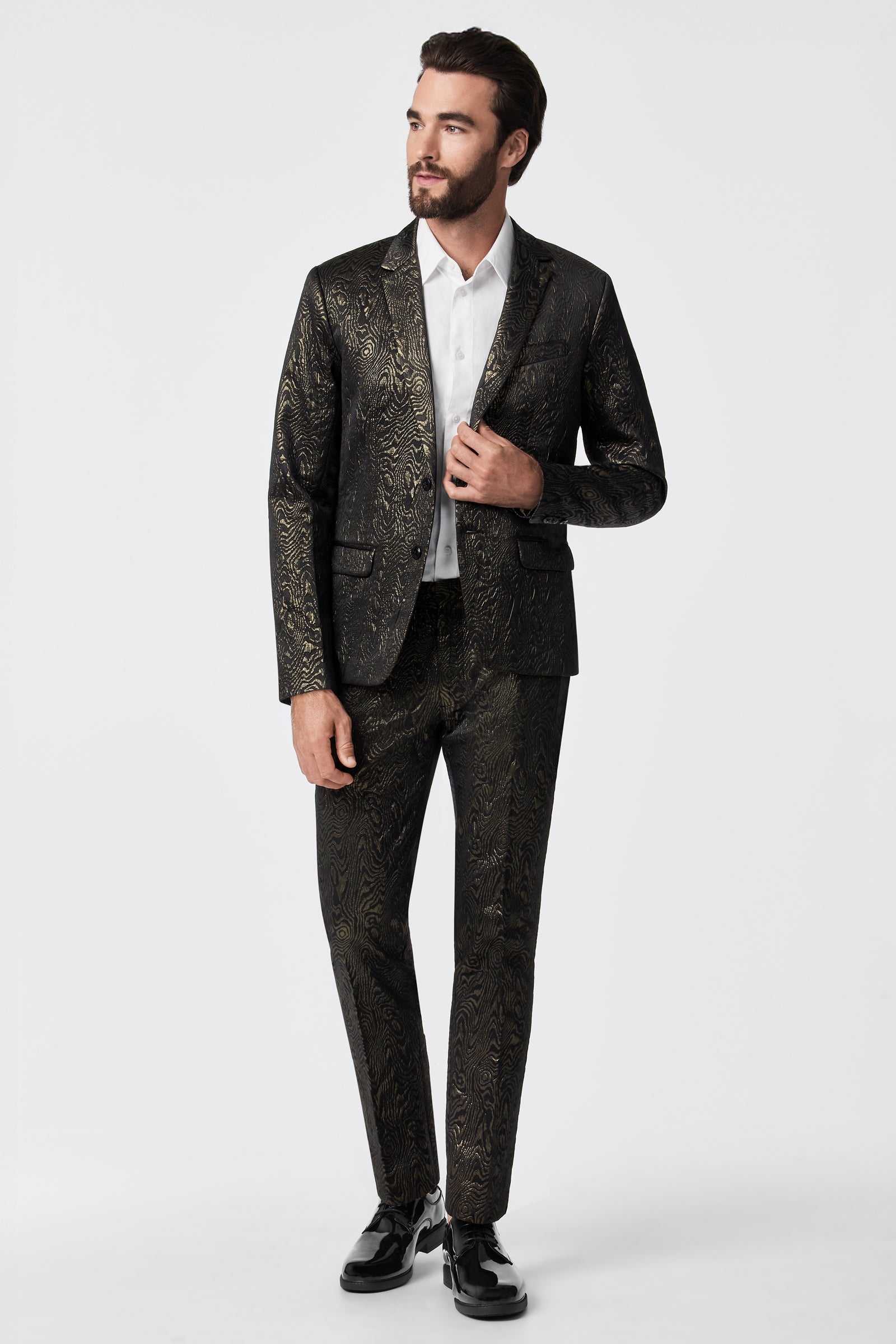 A man with dark hair and a beard wears the Thurston Blazer - Fette Swirl, featuring a black suit jacket with a gold floral pattern, paired with a white dress shirt and black shoes, standing against a plain white background.