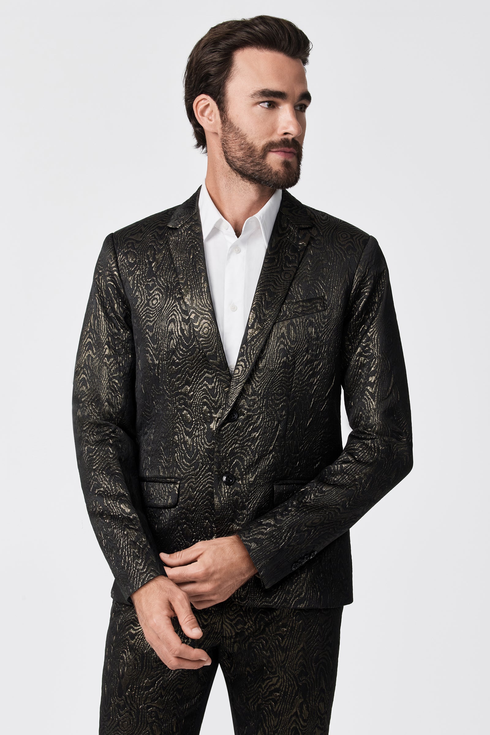 A man with dark hair and a beard wears the Thurston Blazer - Fette Swirl, featuring a black suit with a gold textured pattern over a white shirt. He stands against a plain white background, looking to his left and clasping his hands in front.