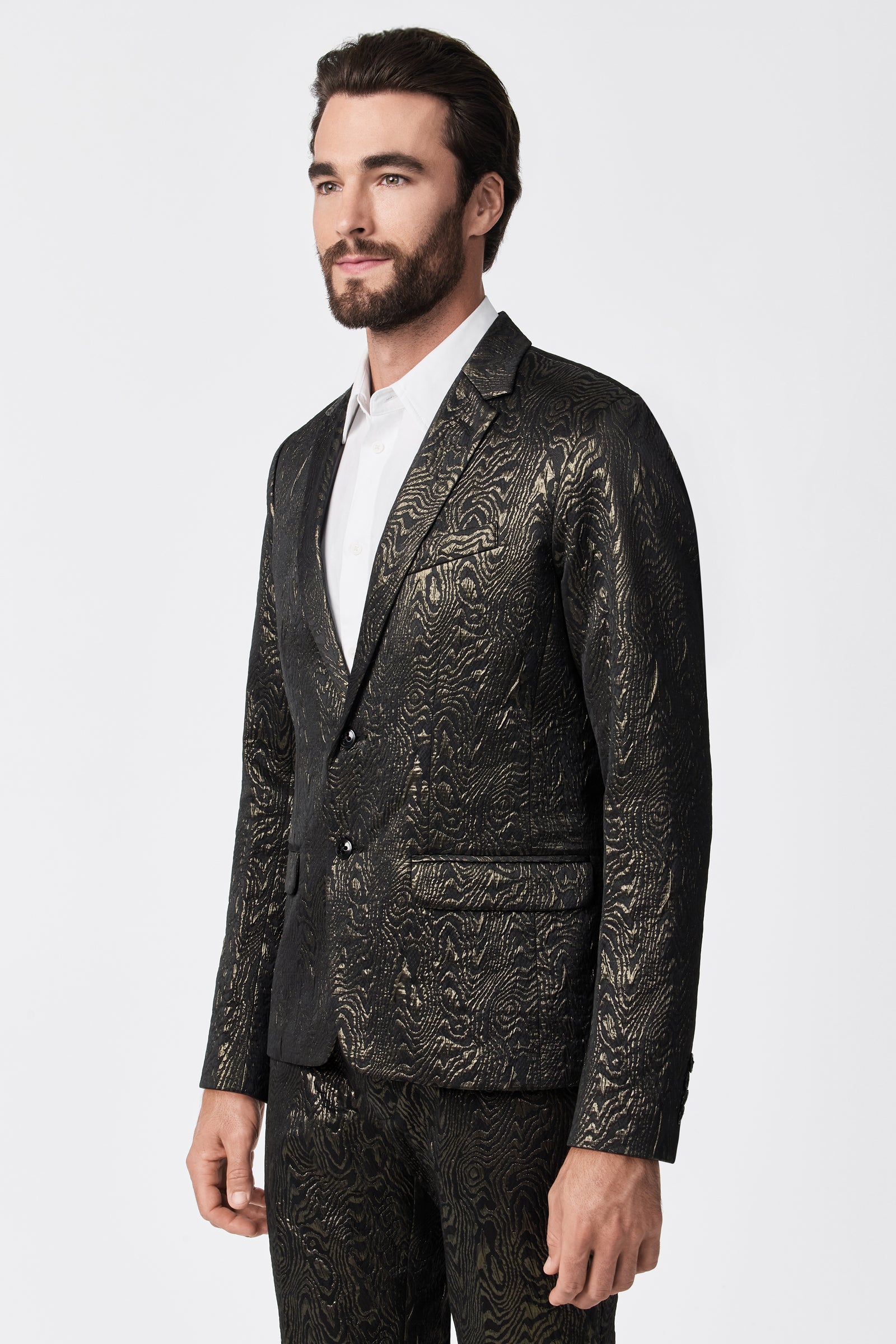 A man wears the Thurston Blazer - Fette Swirl, a dark textured suit with a subtle gold pattern, over a white dress shirt, standing against a plain white background.