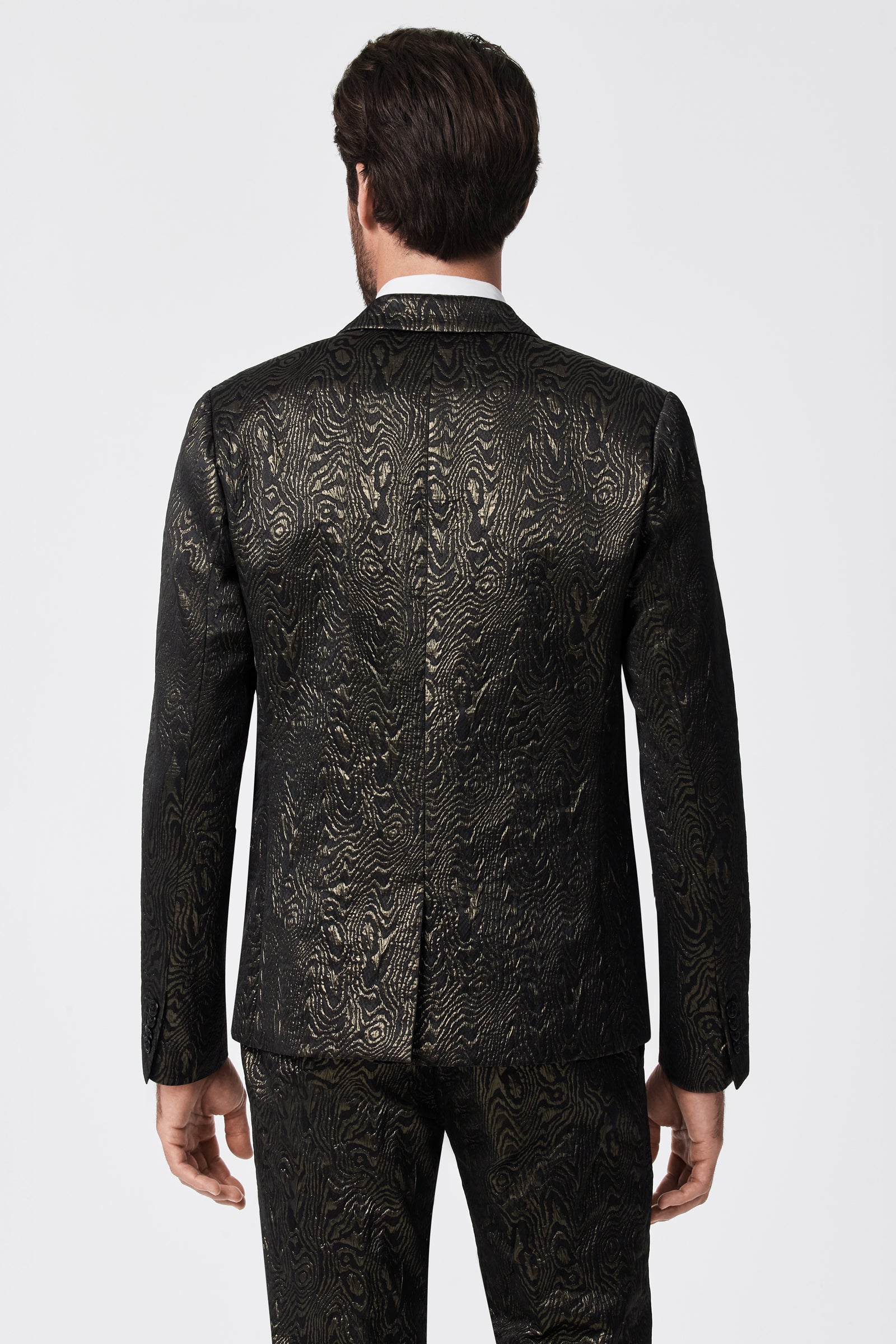 A man stands facing away, wearing the Thurston Blazer - Fette Swirl and matching pants, both featuring a shiny, textured swirl pattern with a metallic sheen, set against a plain white background.