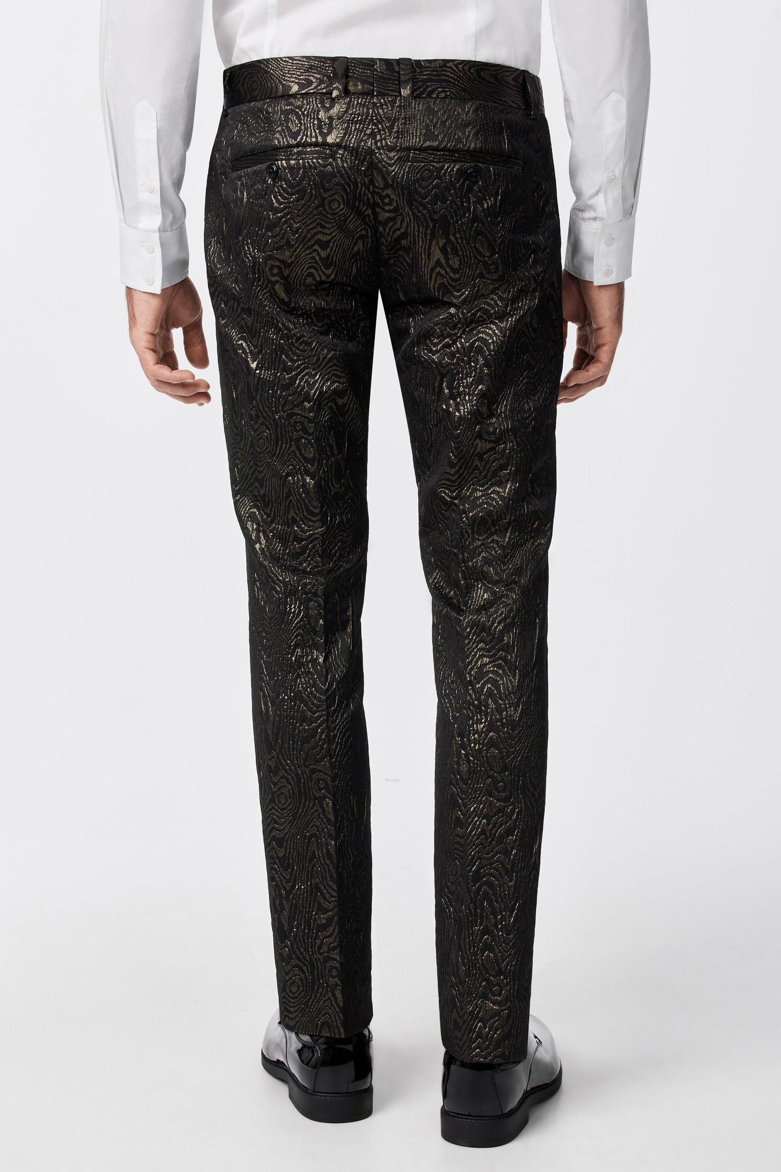 A person is seen from the back wearing the Clyde Slim Trouser - Fette Swirl, featuring a shiny gold pattern, belt loops, and pockets, paired with a white dress shirt and black dress shoes.