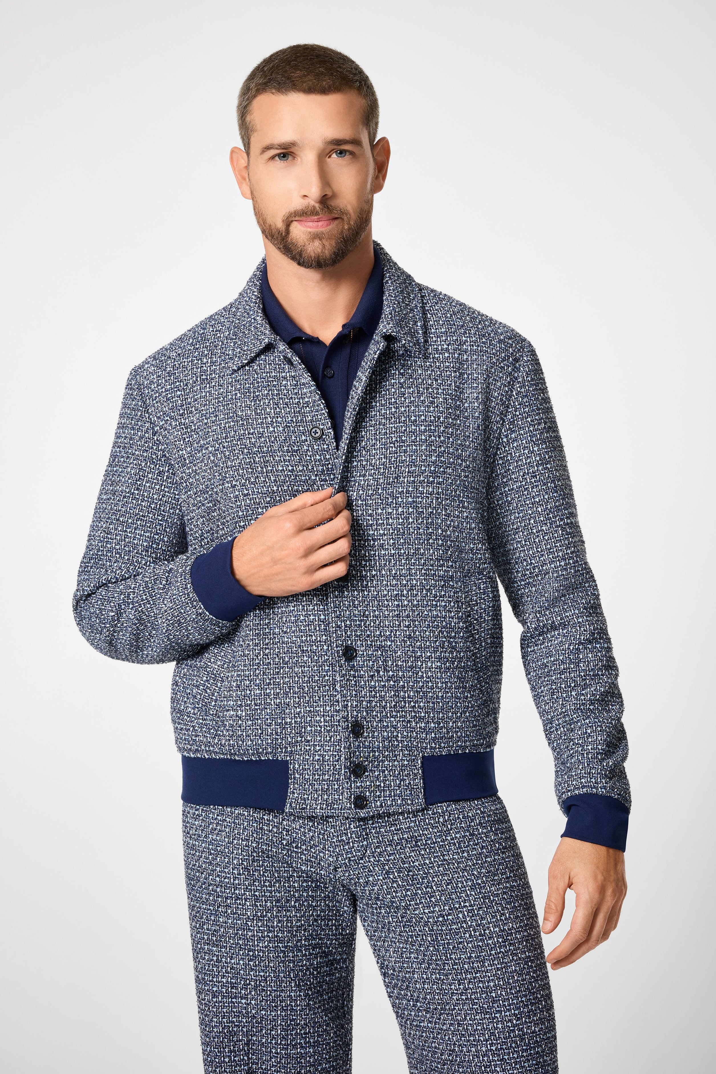 A man with short hair and a beard wears the Lowen Jacket in textured blue with matching trousers and a navy collared shirt, standing against a plain light background.