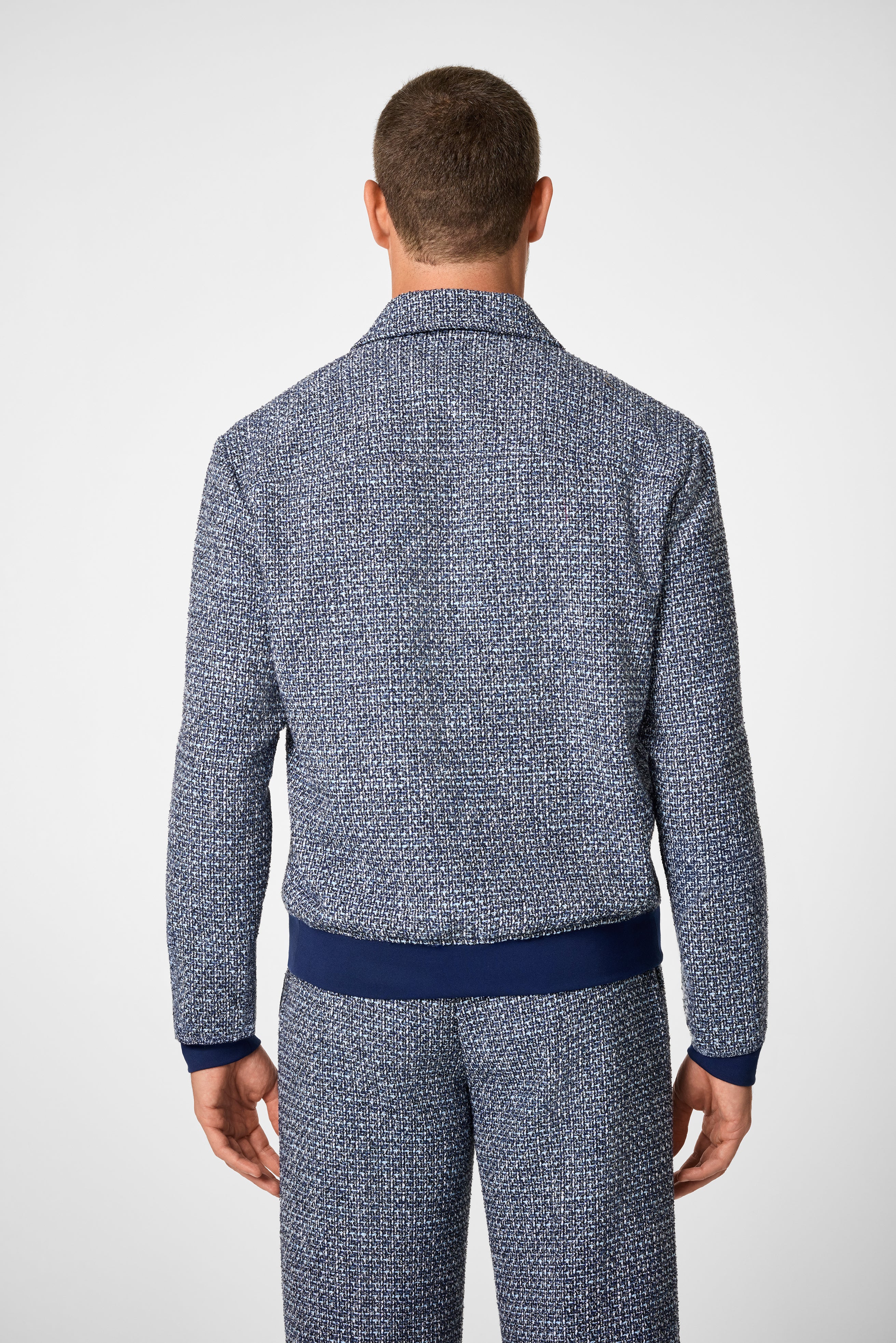 A man stands with his back to the camera, wearing the Lowen Jacket—a blue and white patterned jacket with solid navy blue cuffs and waistband—paired with matching pants against a plain light background.