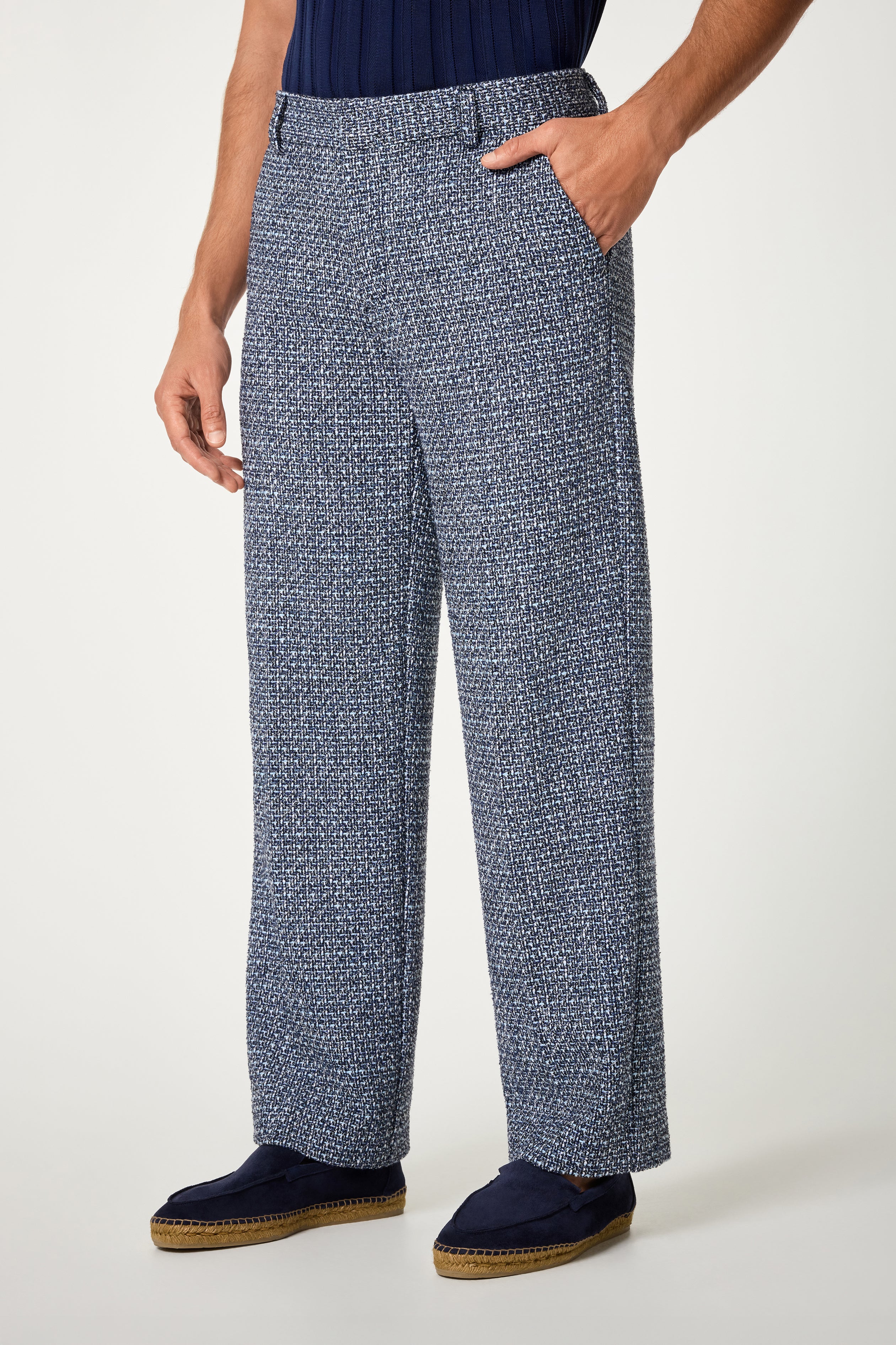 A person wearing the Buckingham Trouser—loose-fitting, textured pants in a blue and white pattern—with hands in pockets, navy slip-on shoes, and a dark blue ribbed shirt against a plain light background.