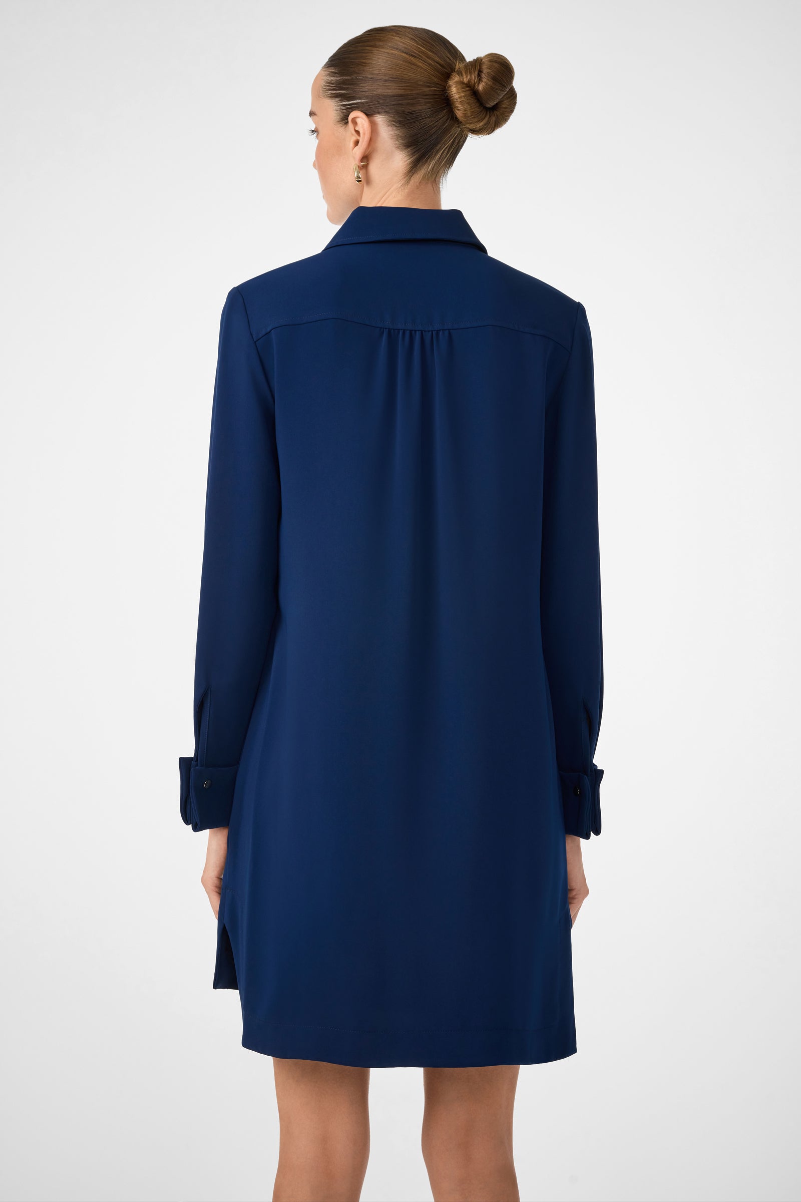 A woman with her hair in a neat bun is seen from behind, wearing the Darya Long-Sleeve Dress in navy blue, featuring a collar and cuffs, standing against a plain white background.