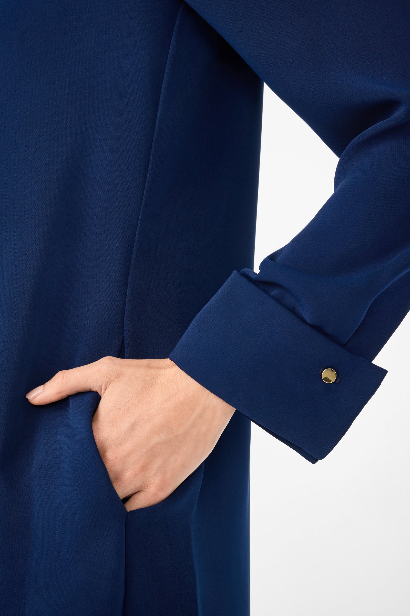Close-up of a hand in the pocket of the Darya Long-Sleeve Dress, a dark blue garment featuring long sleeves with cuffs fastened by small round buttons. The background is plain and light-colored.