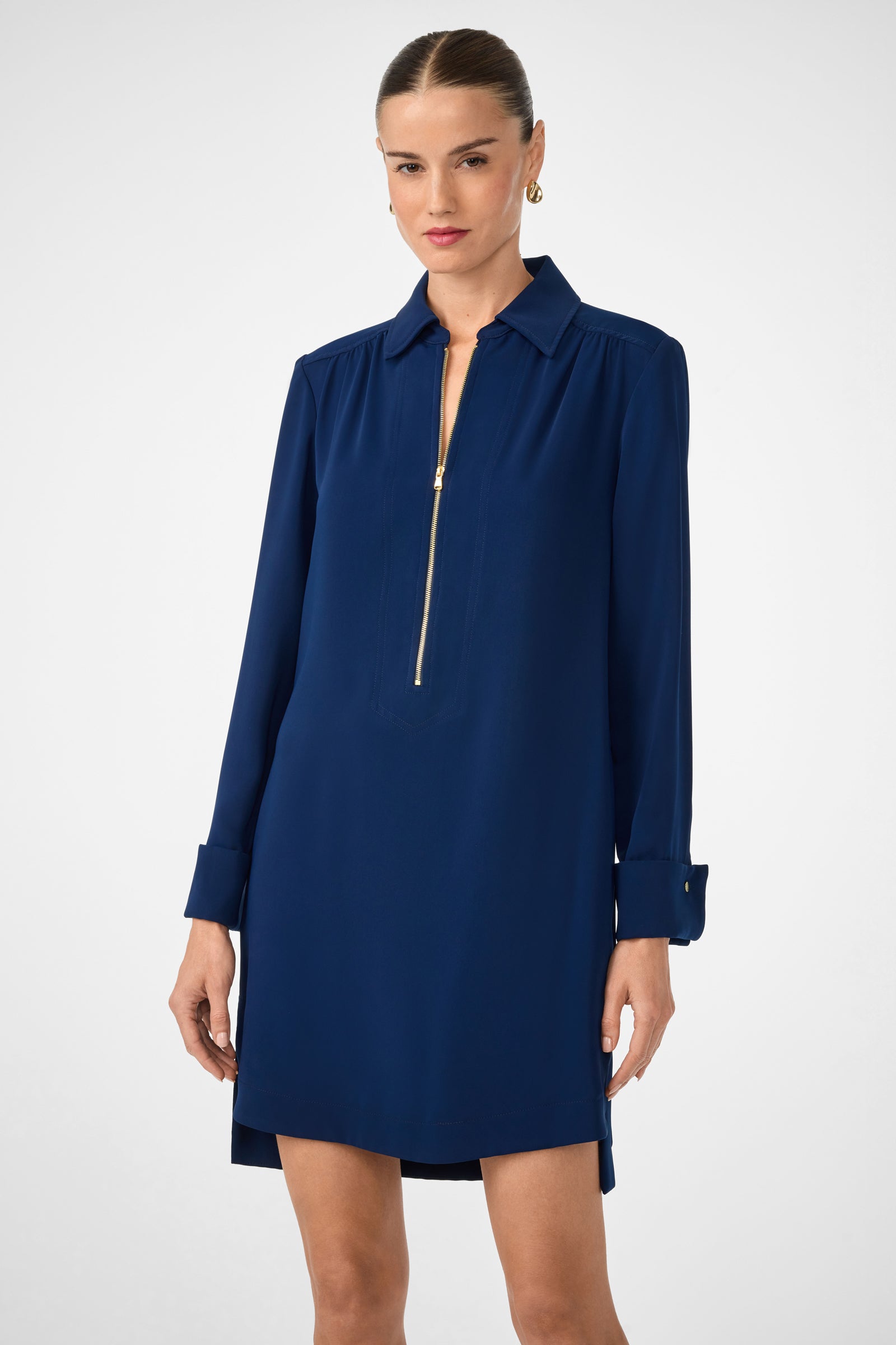A woman stands against a plain light background wearing the Darya Long-Sleeve Dress—a navy blue shirt dress featuring a collar and front gold zipper. Her hair is pulled back, and she accessorizes with small hoop earrings.